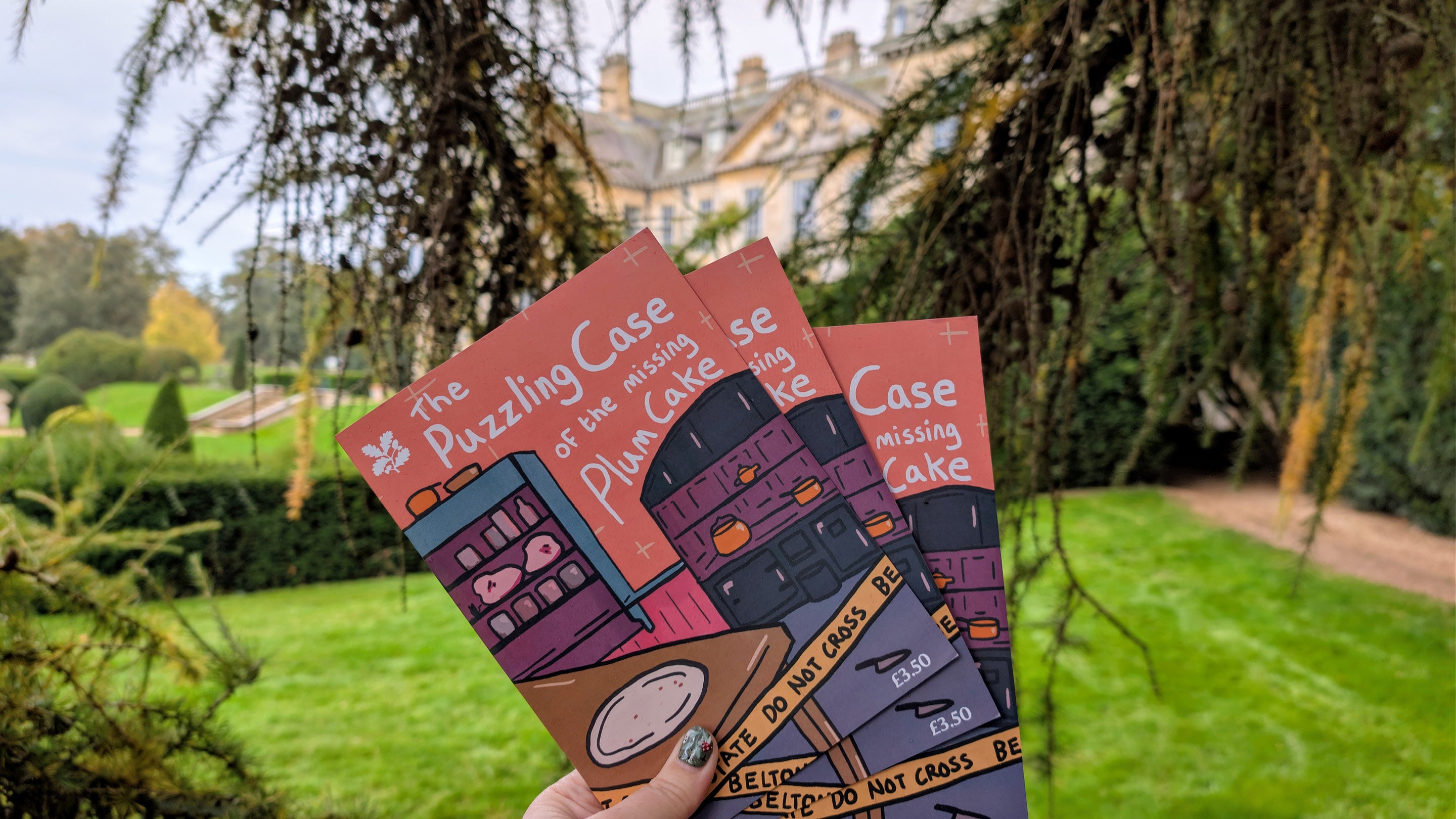 Three copies of The Puzzling Case of the Missing Plum Cake mystery guide are held in the gardens with Belton House behind through trees.