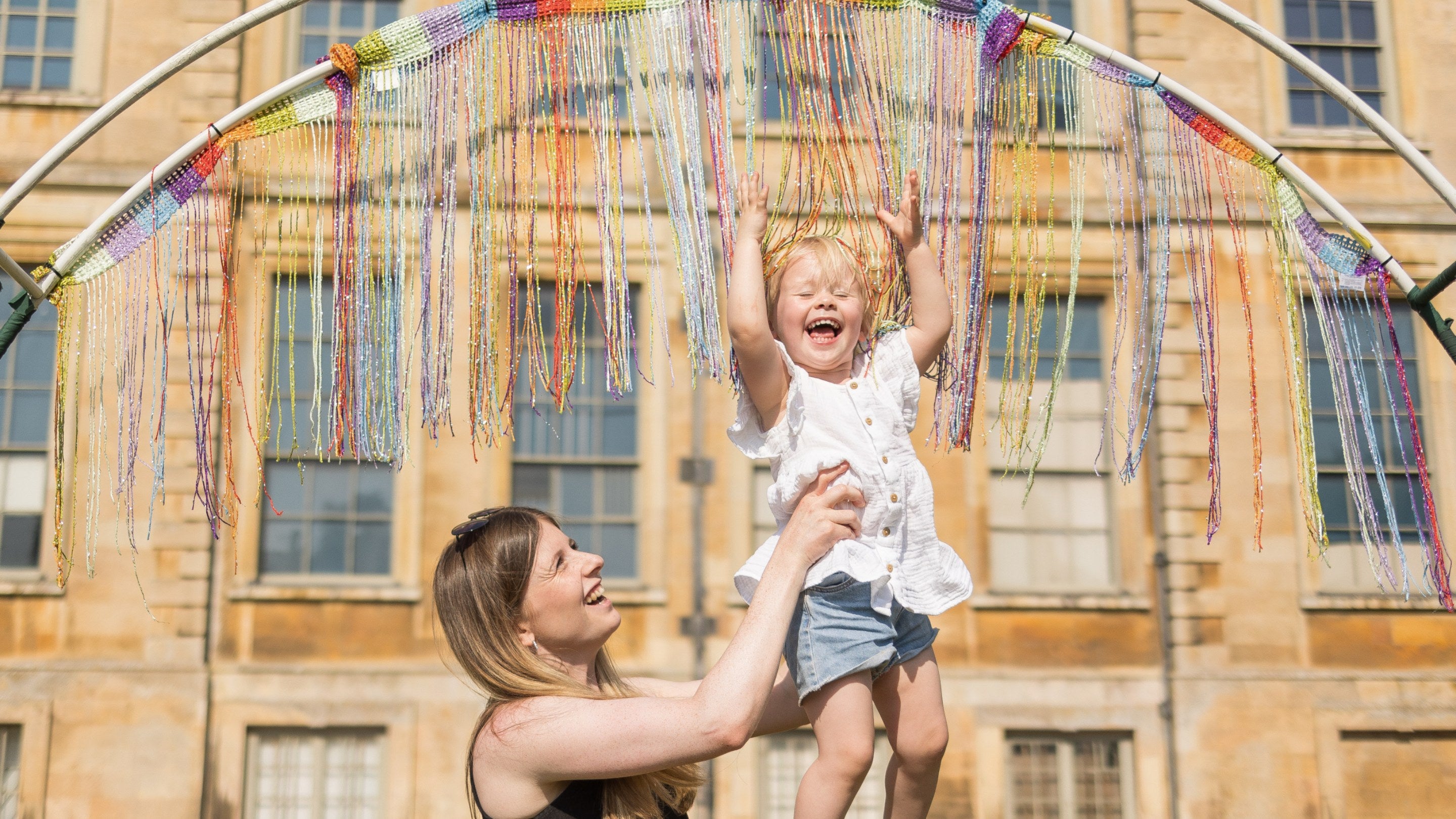 A woman holding up a young child so that their head is touching the multicoloured strings that are falling from the top of a semi-circled piece of metal. Both parent and child have huge grins on their faces.