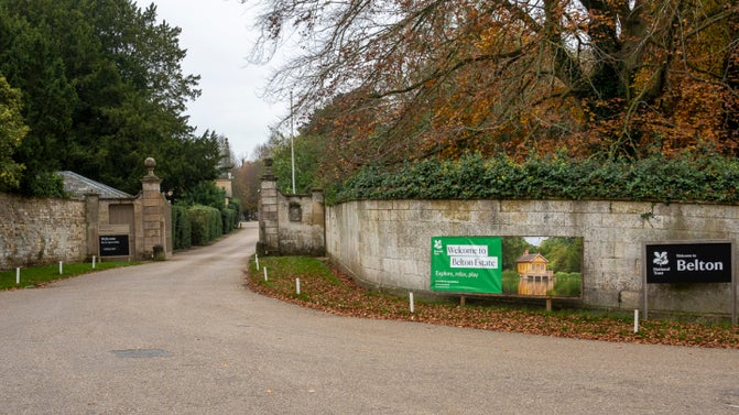 The main entrance to Belton Estate. The road curves into an open gate between two stone walls. There is a black Welcome to Belton Estate sign on the right.