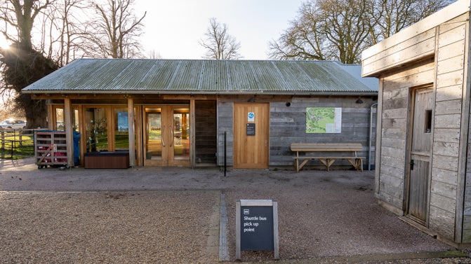 Belton's Information and Accessibility Hub. A wooden building with a grey corrugated roof, wooden doors and supports. There is a fold out A-board on display in the foreground which says shuttle bus pick up point..