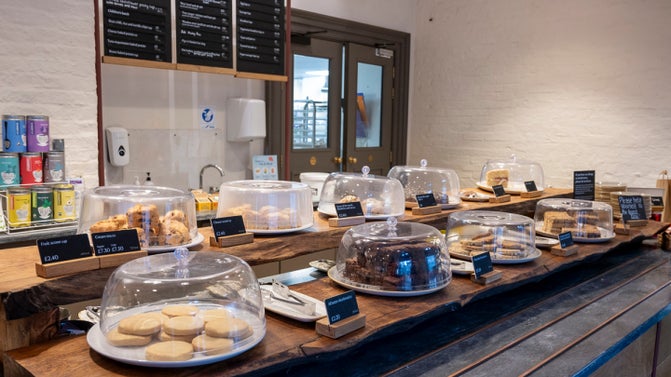 The servery at the Stables Café with an array of cakes under plastic domes and black menu boards hanging in the background.