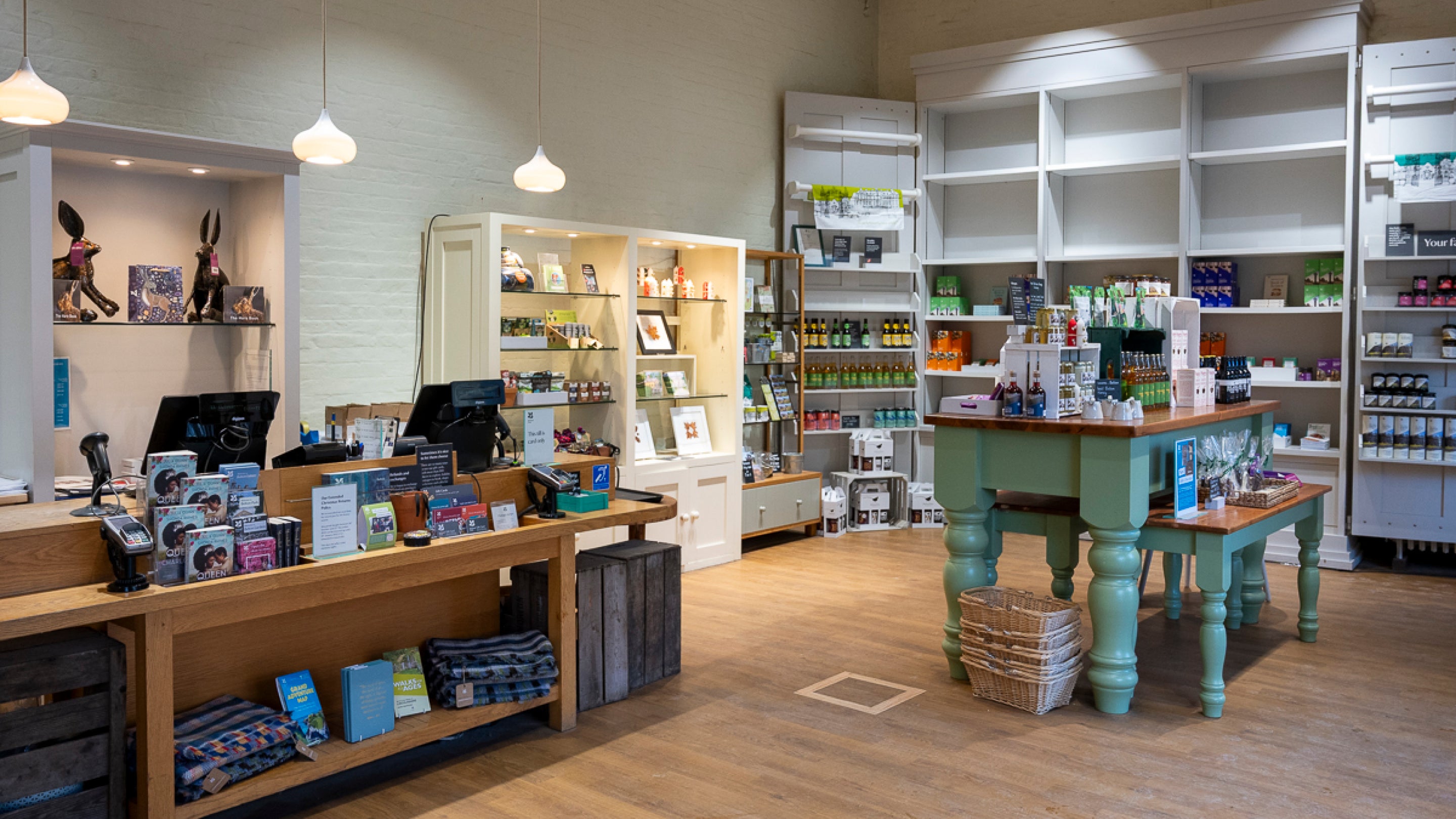 The Old Coach House Shop with shelves of products against the walls and a large green display table. To the left of the image is the till point which is a wooden desk with two tills, decorated with leaflets and products.
