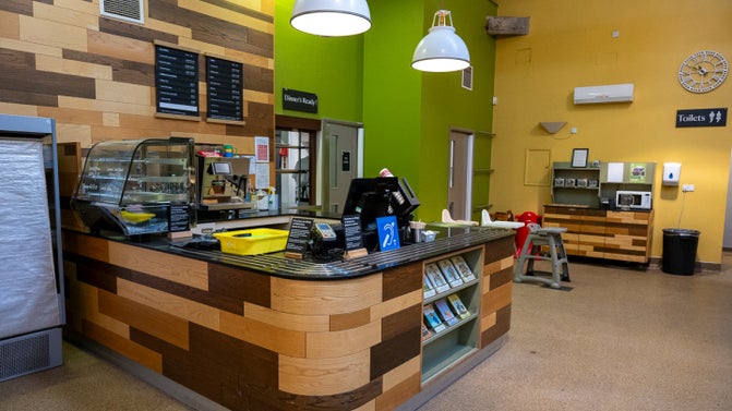 The servery in The Ride indoor play café. The till point is made of different colours of wood with a black counter. There is a fridge unit to the far left and a hot food display.
