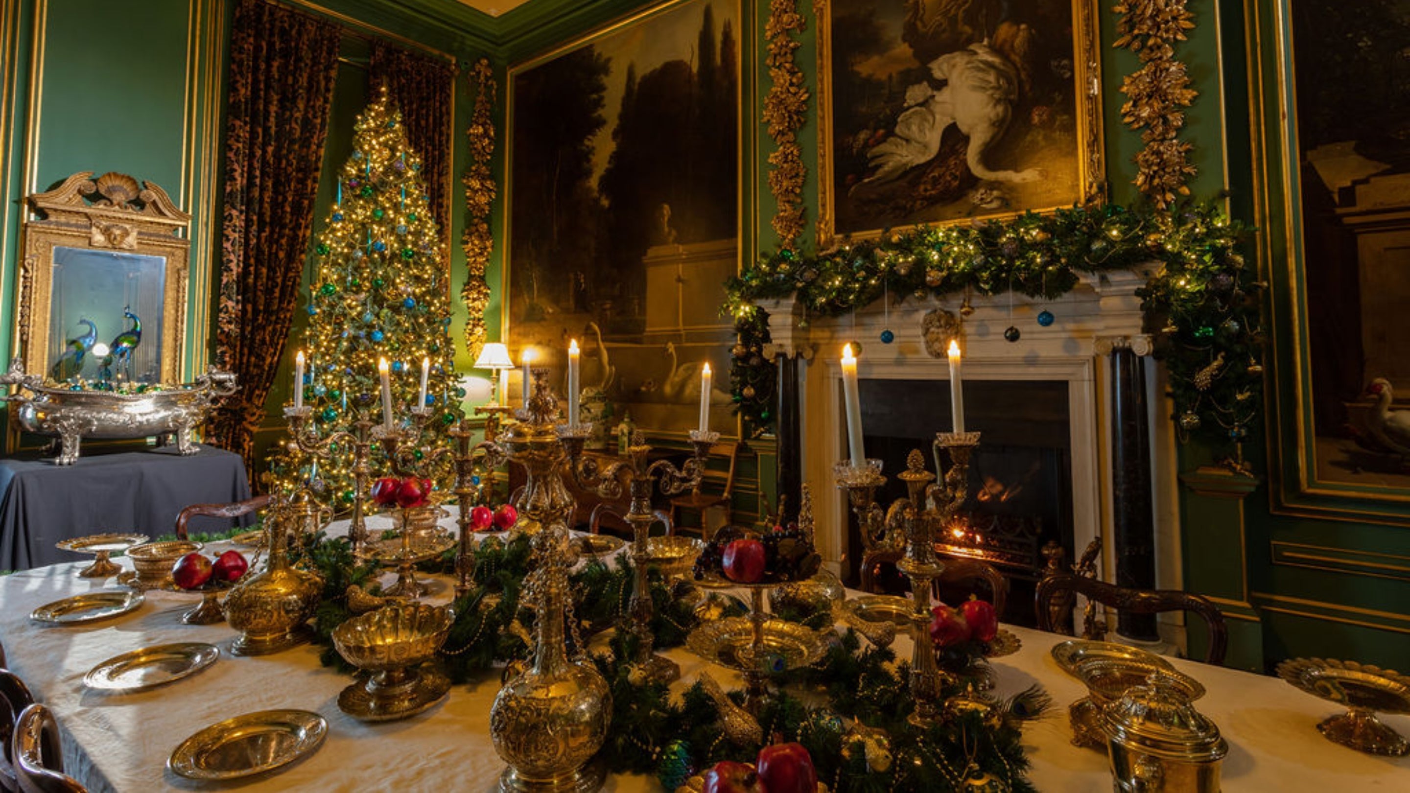 Image showing Christmas decorations inside the Hondecoeter dining room at Belton, with large tree in the corner dressed in blue and green baubles. The table has a silver gilt collection with a decorated green garland, with one to match on the mantlepiece.