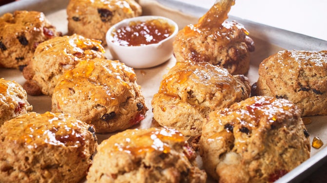 Several freshly baked fruit scones on a baking tray. The scones have had an amber coloured jam applied to the top of them. Also on the baking tray is a small white ceramic pot containing amber coloured jam. The tip of a brush can be seen applying jam to one of the scones.