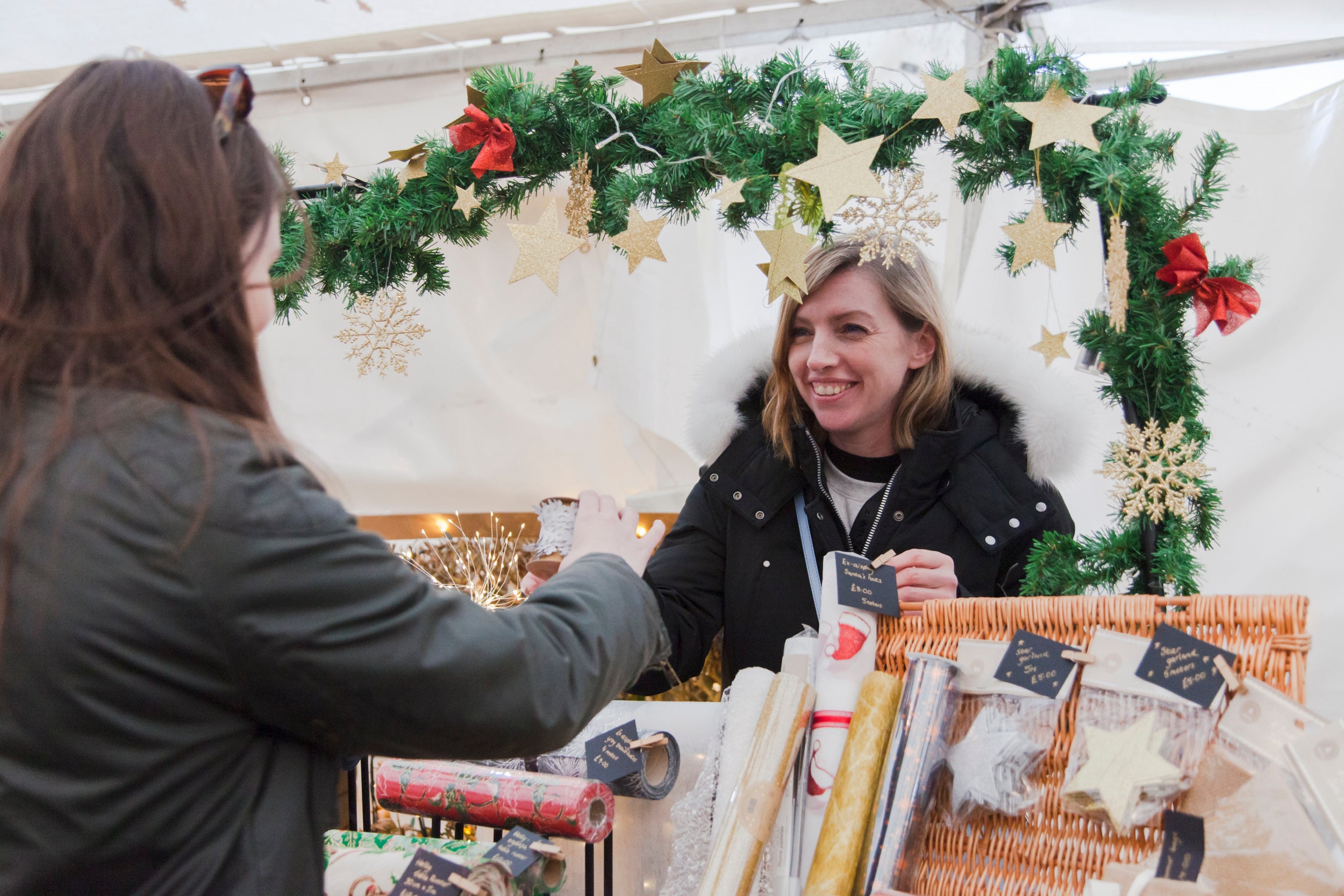 A woman smiles as she serves a customer from behind the counter of a Christmas Market stall. The stall is decorated with a festive garland and there is a wicker basket with wrapping paper and gift tags for sale.
