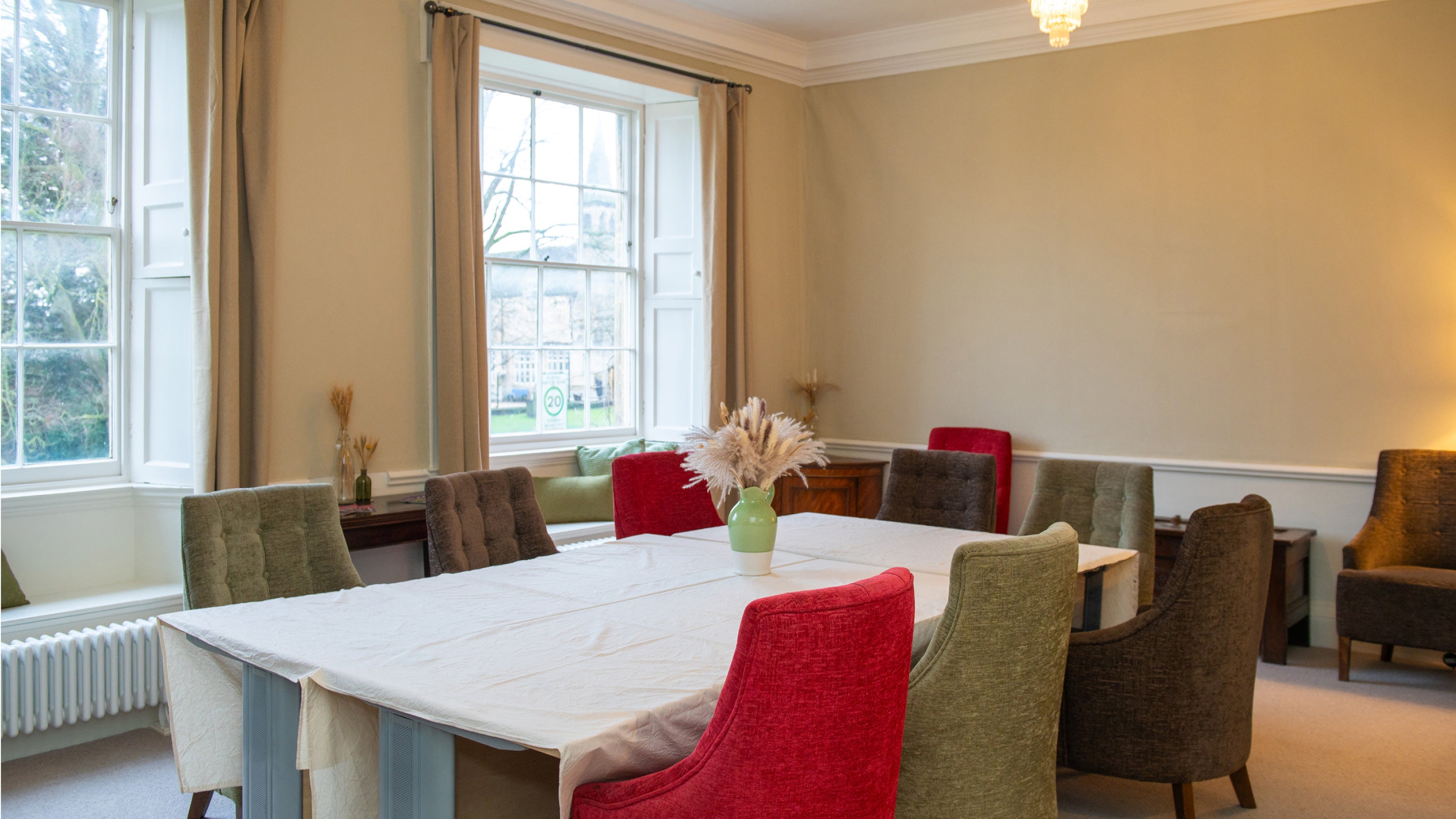 The Sedgwick Room at Grantham House. It has pale green walls, two large sash windows with a view of the lawns and the neighbouring St Wulfram's Church, and a large table with white tablecloth and red and green chairs.