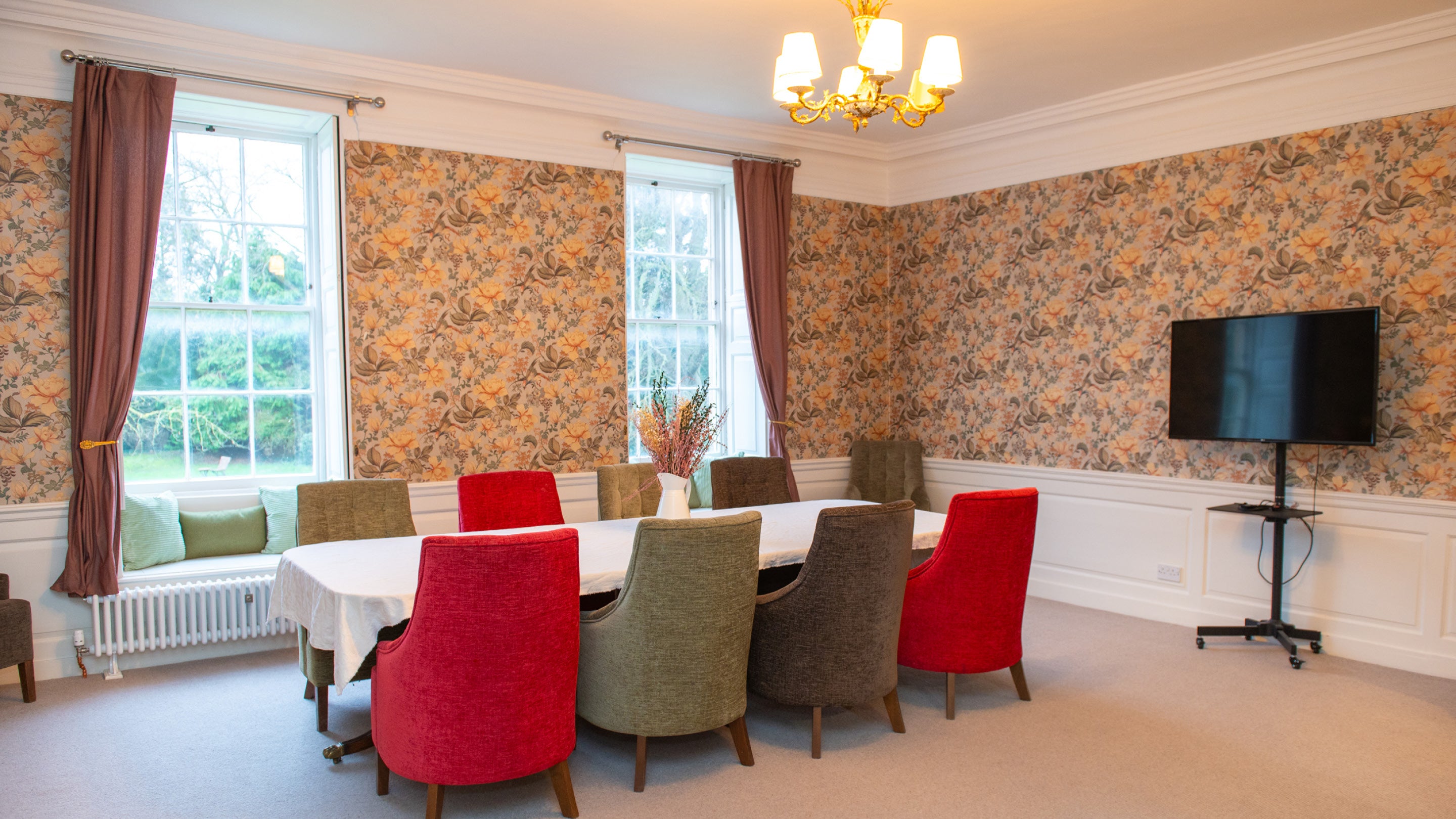 The Wyldbore Room at Grantham House. The wallpaper is orange and green with a floral design, there are two large sash windows and a long table set with red, green and brown chairs.