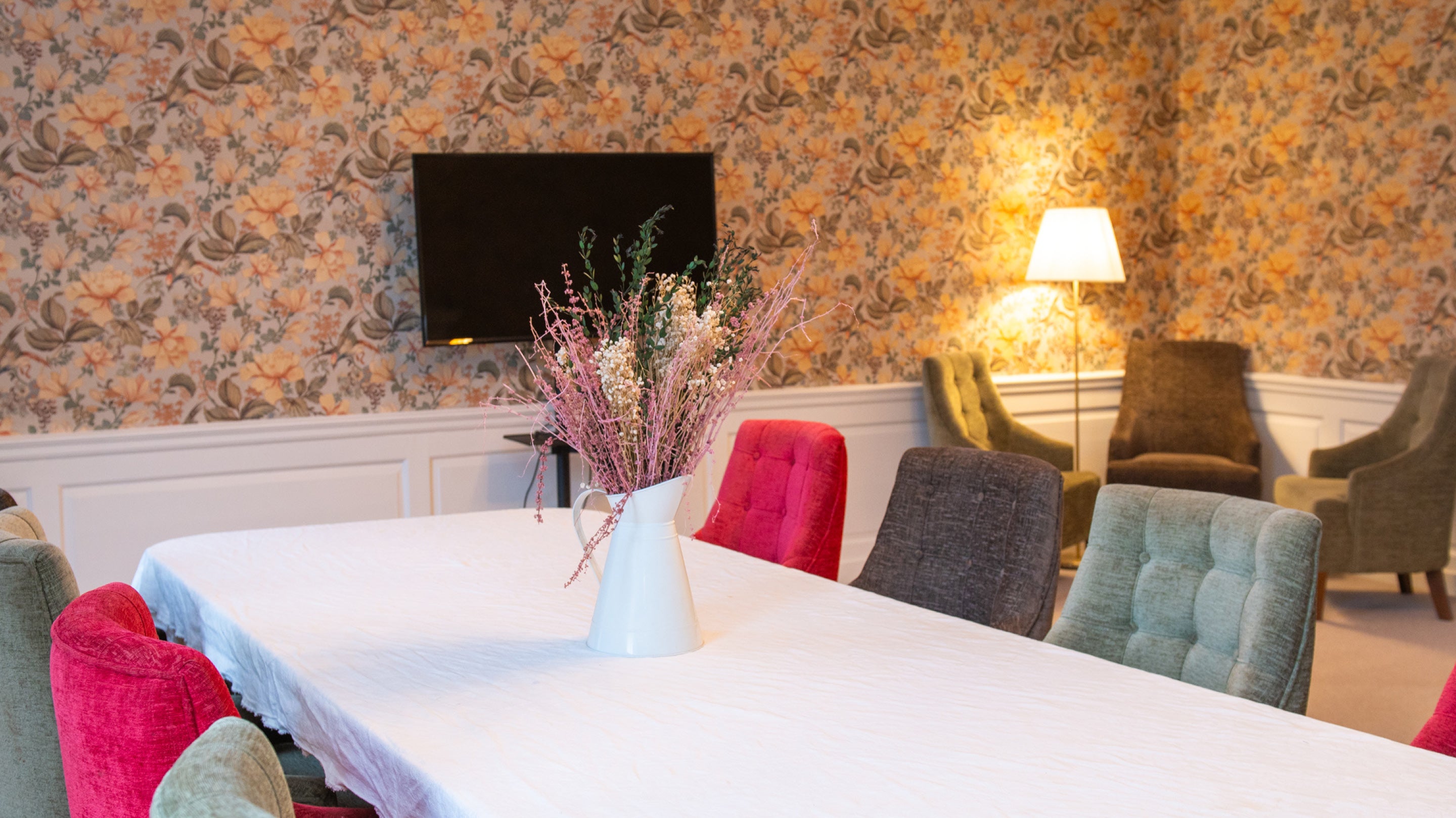 A white table with red, green and brown chairs in a room with orange and green floral wallpaper. A TV screen is mounted on the wall and three green chairs are in a cluster in the corner around a table and lamp.