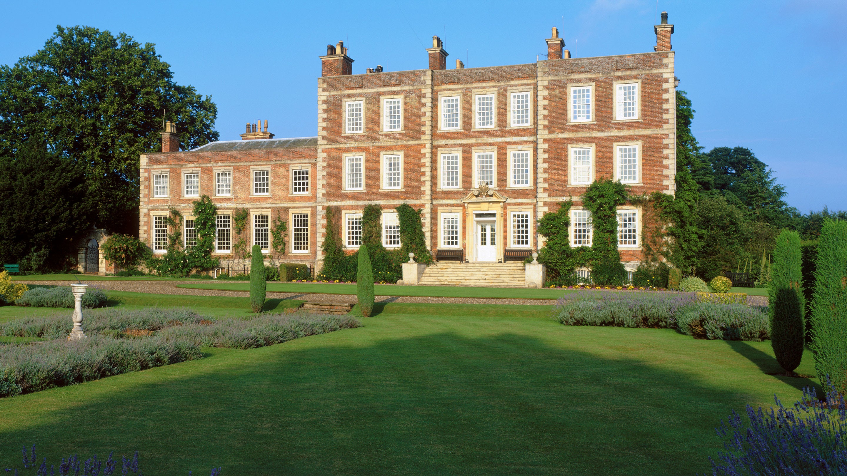 View of the front of Gunby Hall showing lawns and lavender beds with a sundial. The house is basking in the evening sunlight.