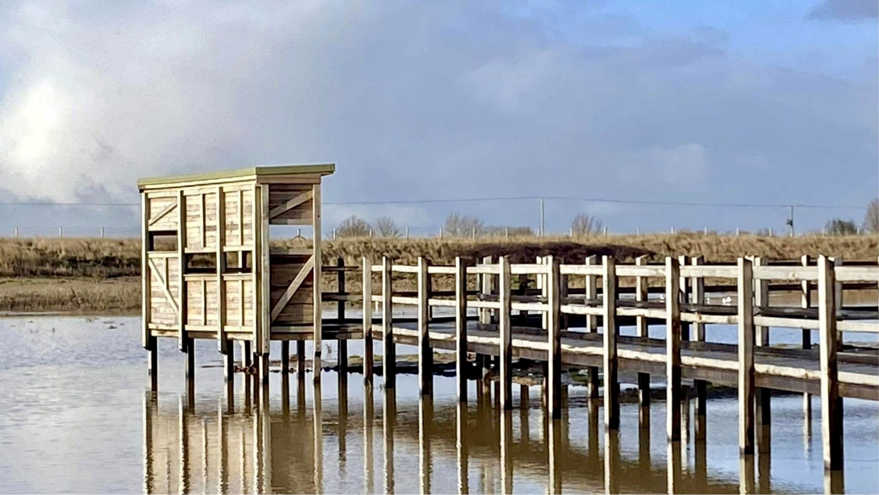 The wooden boardwalk leading to a small bird screen over shallow wetlands at Sandilands Nature Reserve, with reflections in calm water under a cloudy sky.