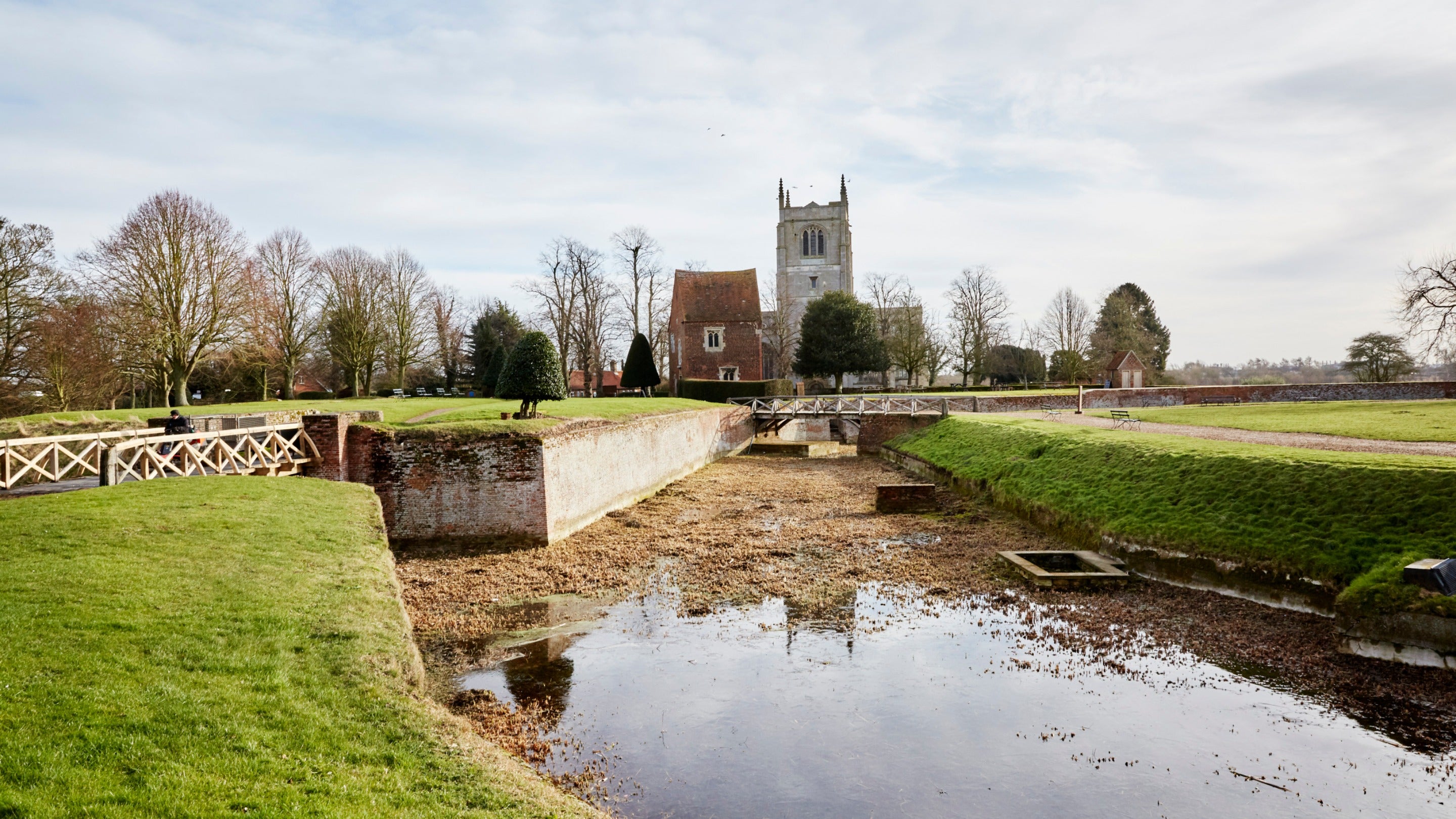 A view of the moat at Tattershall Castle, with two small bridges crossing the moat and a view of the Guardhouse and Church of the Holy Trinity in the background