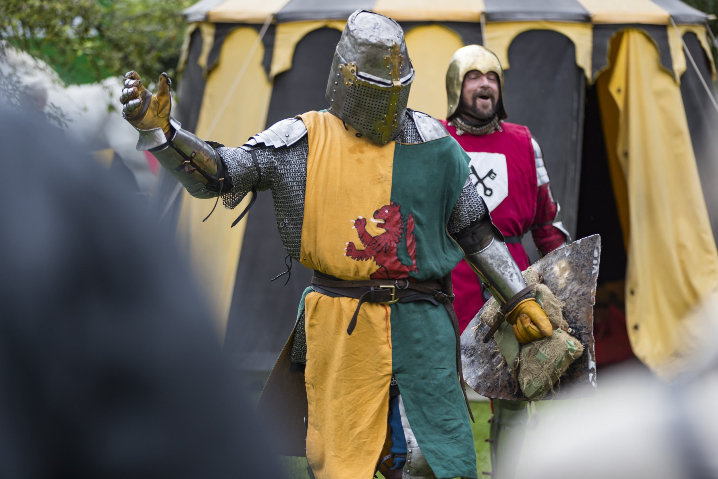 A medieval knight with a helmet on holding his arm in a victory pose while another knight laughs behind him. There are tents in the background.