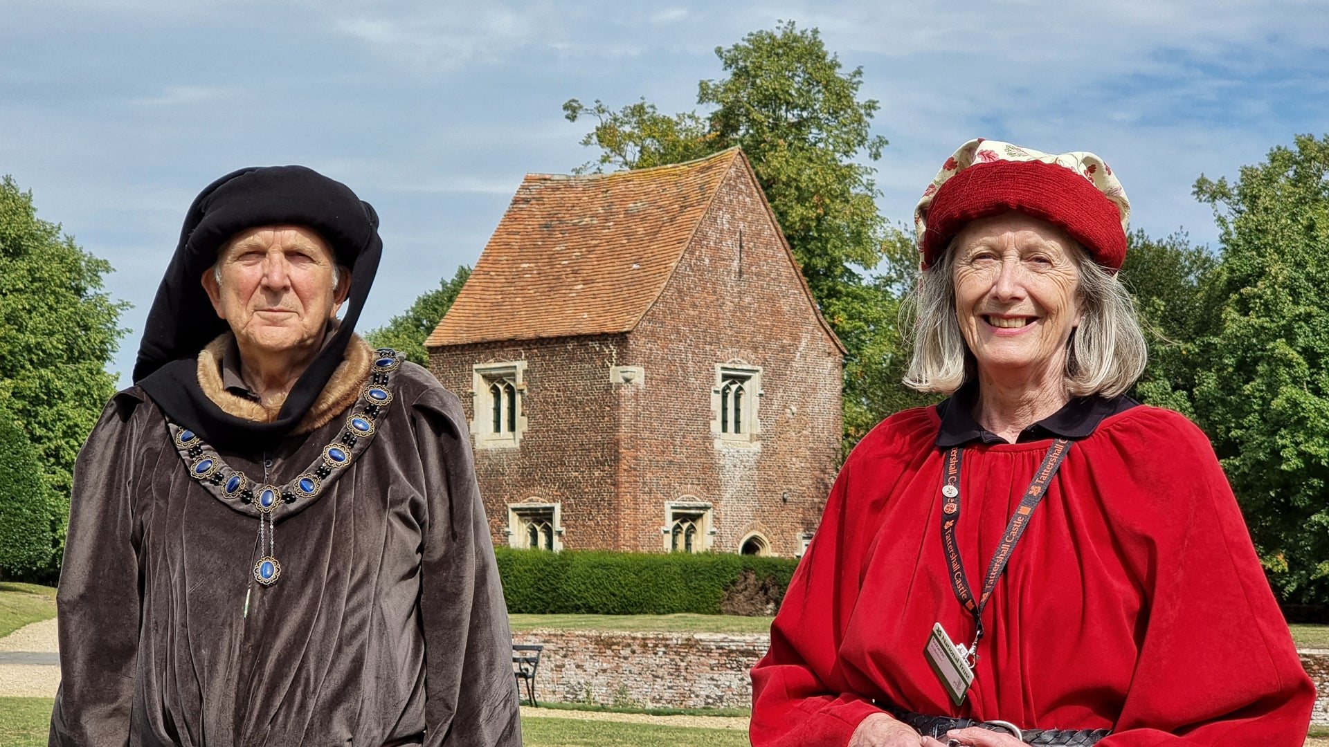 Two volunteers at Tattershall Castle wearing medieval costumes. The visitor reception building is seen behind them.