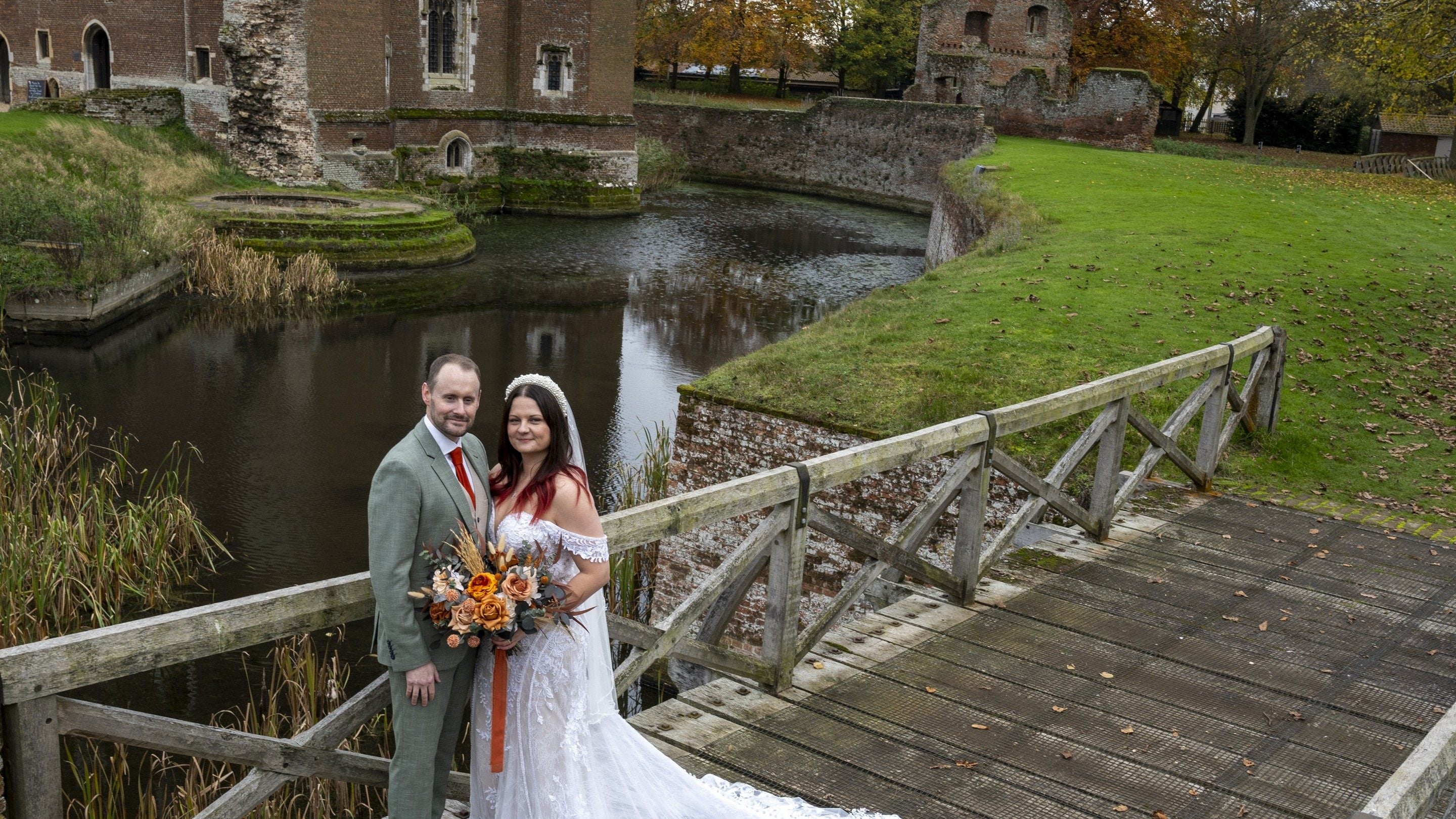 Couple in wedding attire standing on a wooden bridge over a moat at the historic brick, Tattershall Castle, with autumn trees and grassy grounds surrounding the water. Lincolnshire wedding venue.