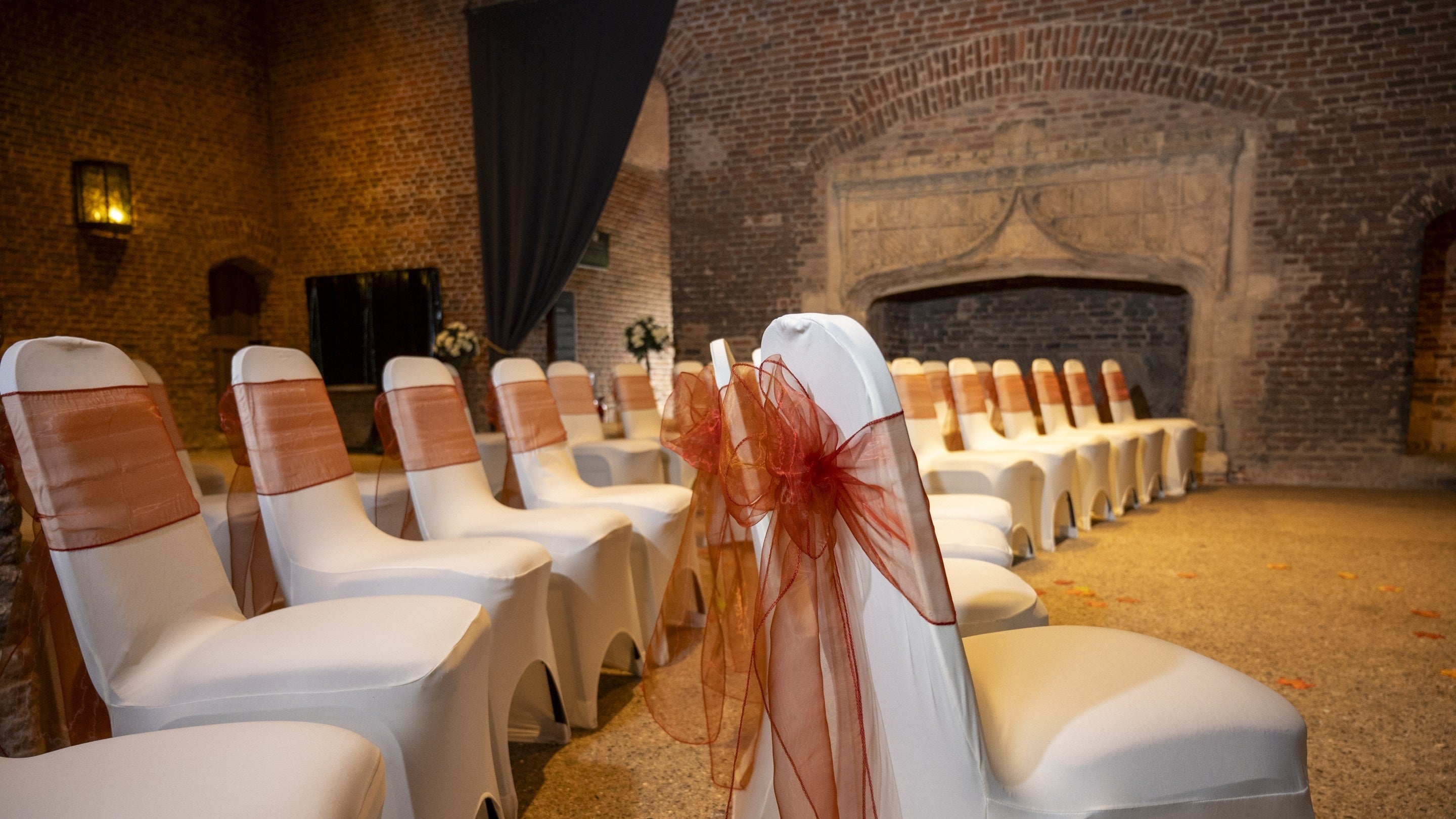 Rows of white-covered chairs with burgundy bows arranged for a wedding ceremony in the historic Parlour at Tattershall Castle, with medieval brick walls and an ornate stone fireplace in the background. Lincolnshire wedding venue.