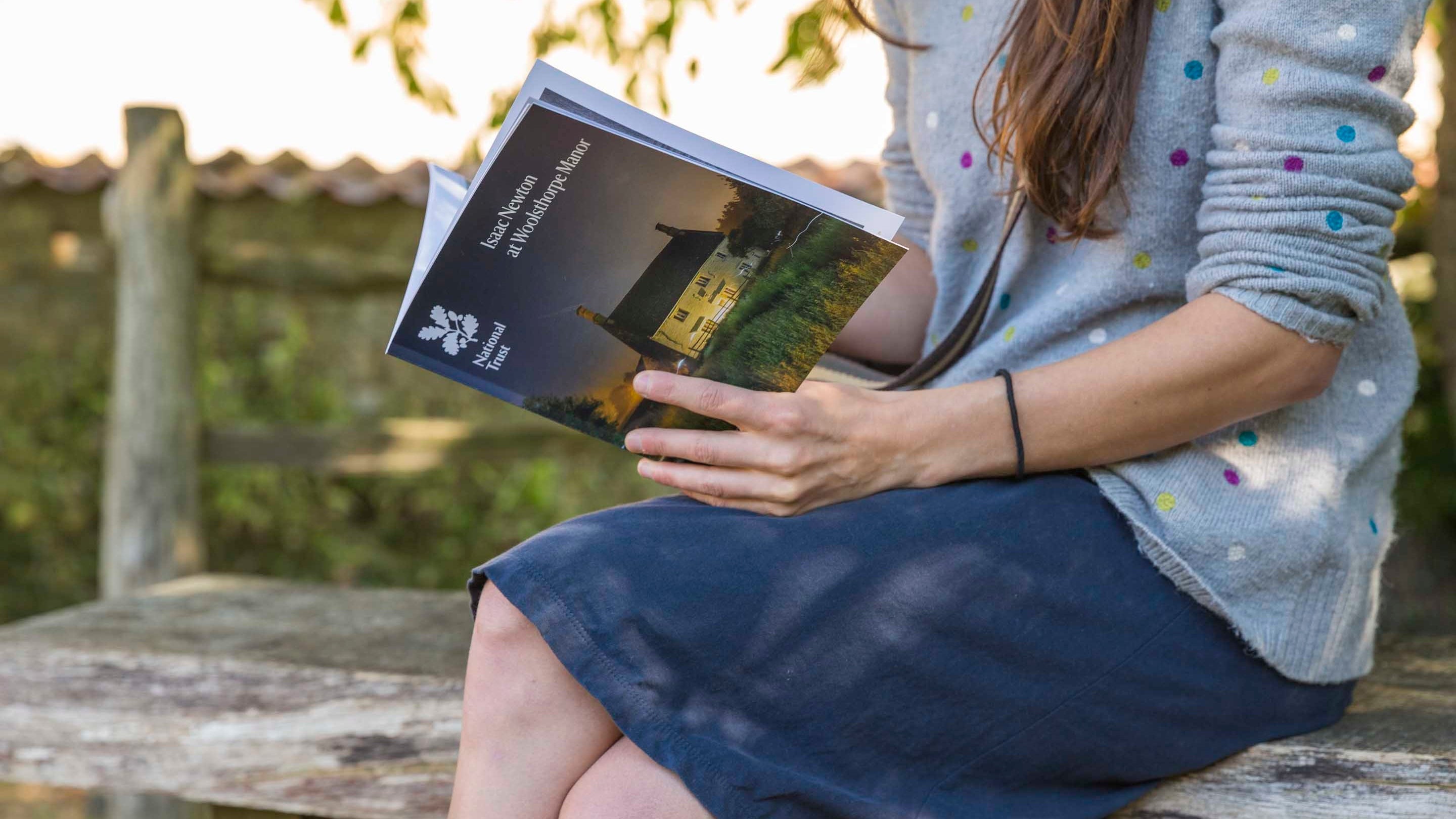 A visitor sits on a bench in the garden reading the Woolsthorpe Manor guidebook.