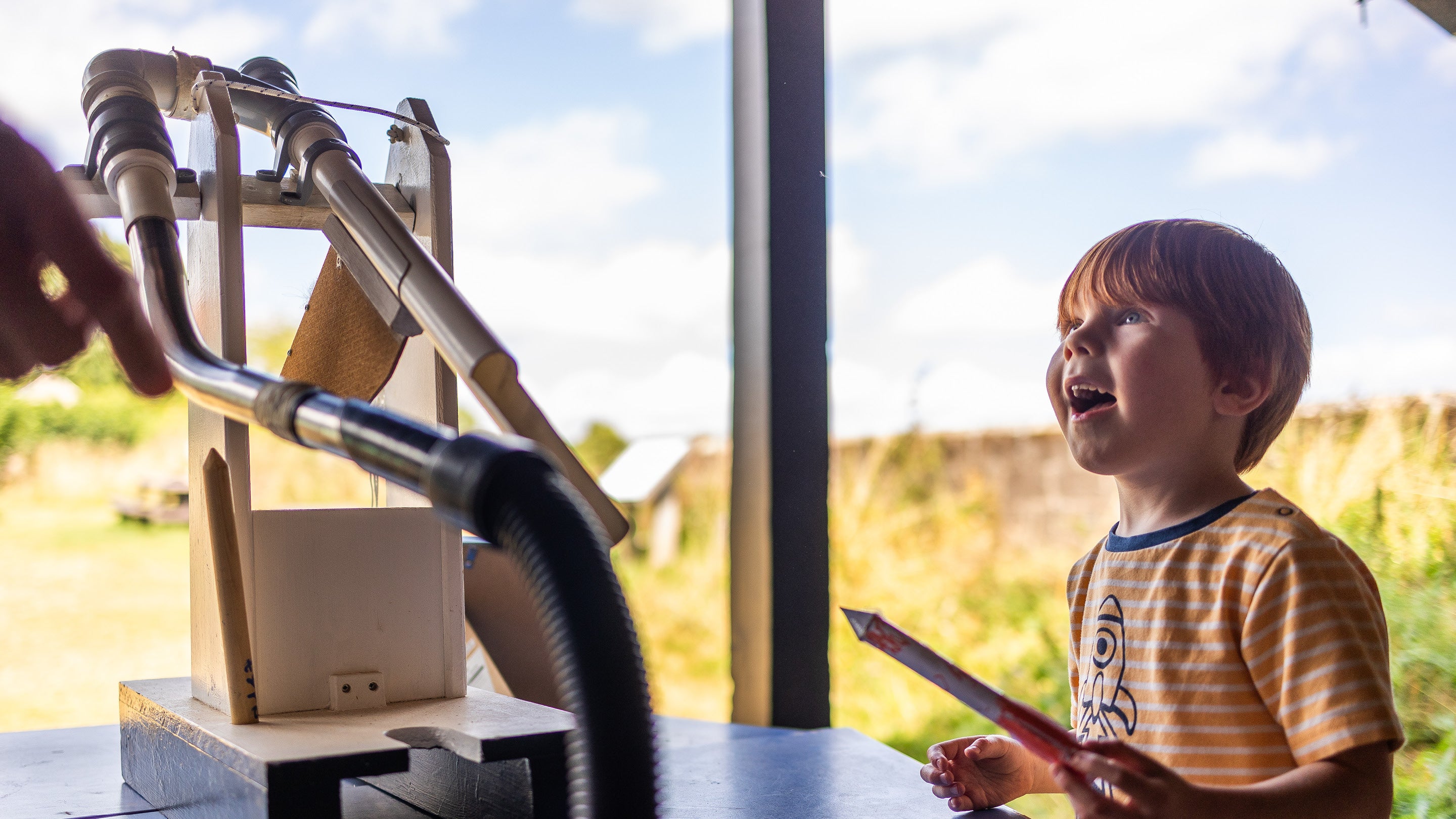 A young boy smiles while holding a paper rocket, ready to launch it using a contraption next to him.