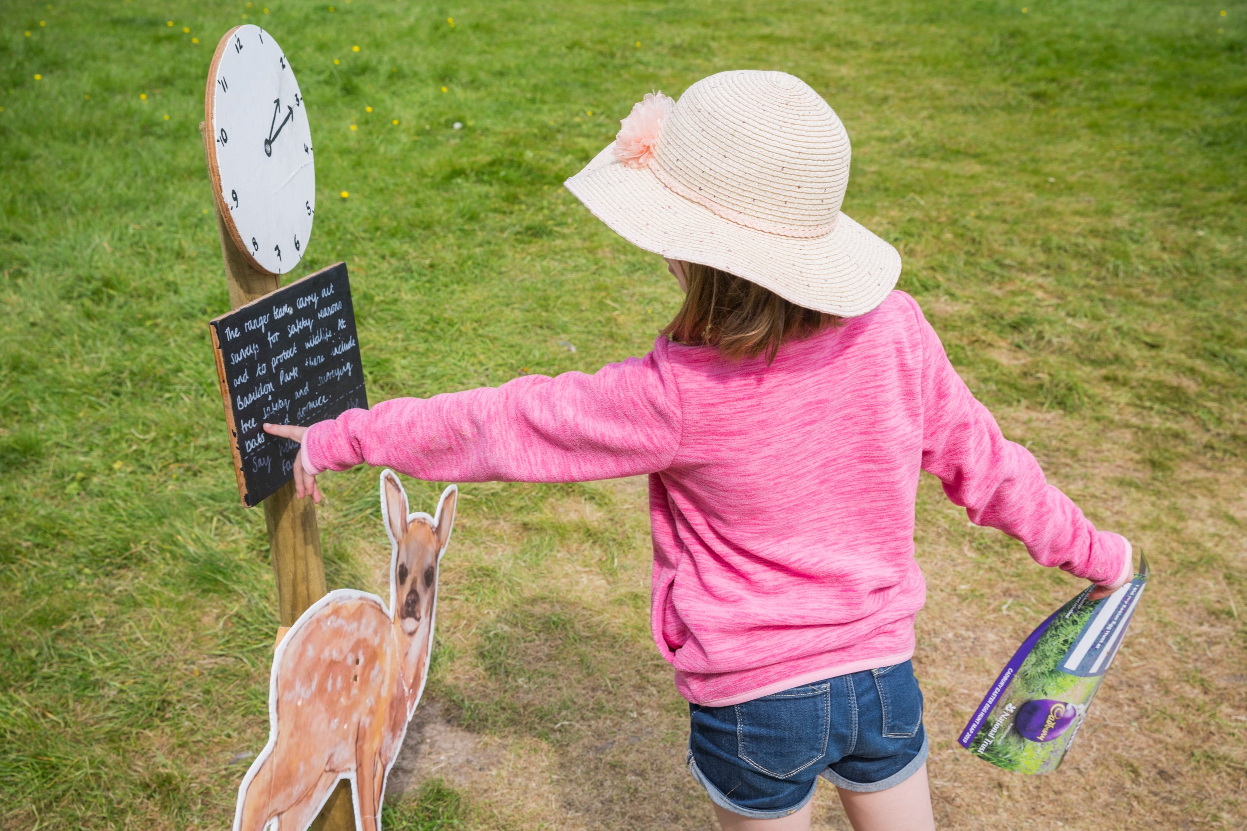 A child wearing a straw hat and a pink top is point at a clock as part of an Easter trail