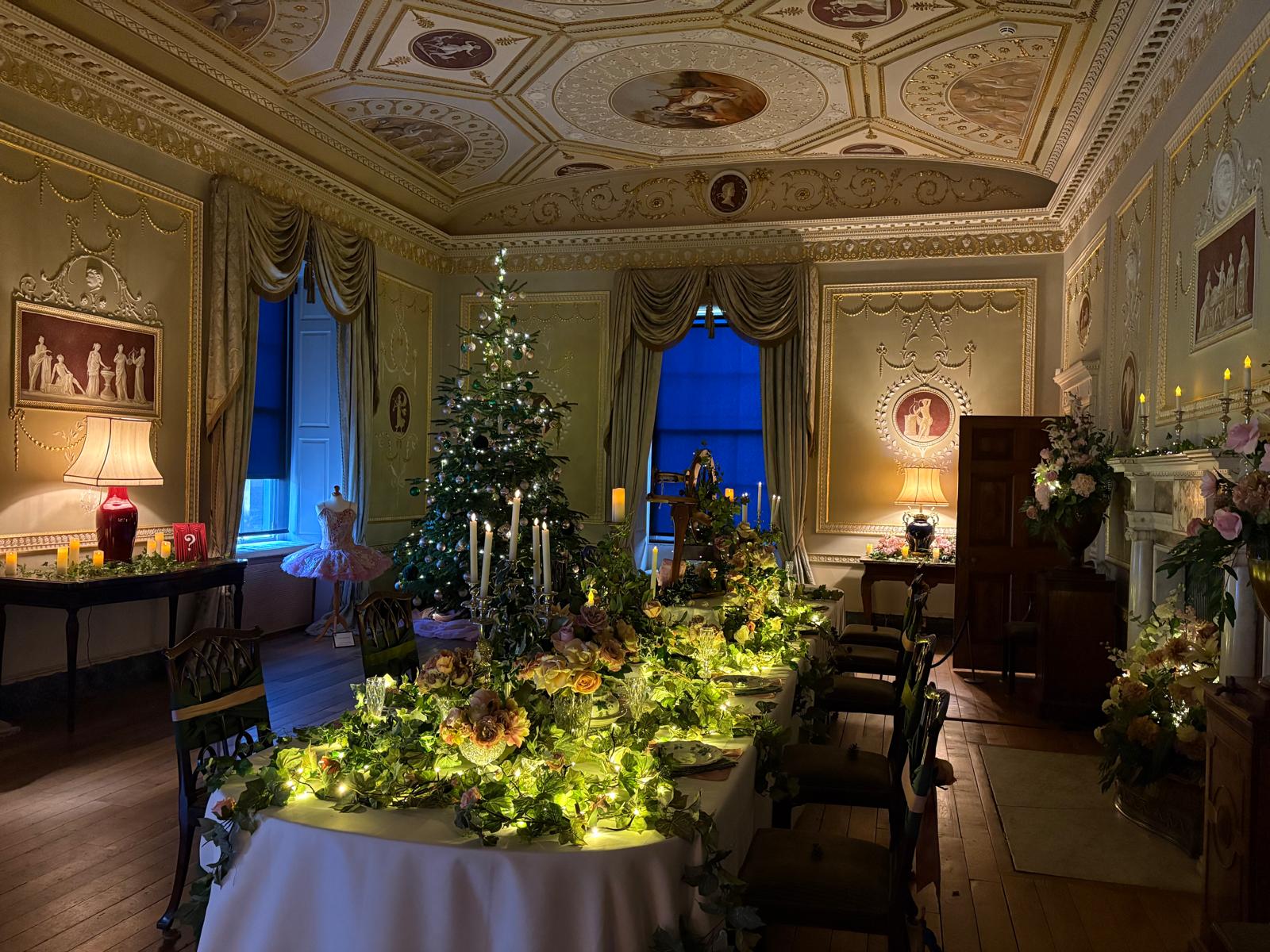 Dining room decorated for Christmas with a dining table covered in ivy, pink flowers and candlesticks. In the background is a Christmas tree decorated in green and pink baubles with ivy.
