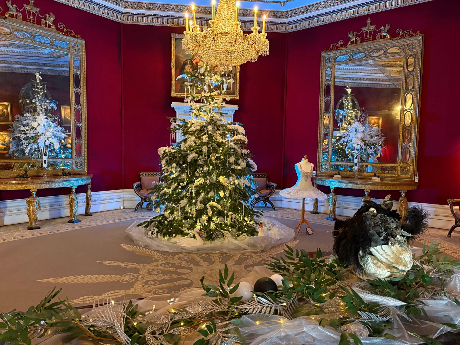 A red octagonal room decorated for Christmas. In the centre is a Christmas tree decorated with white feathers and baubles. Next to the Christmas tree is a white tutu costume. Around the room are white floral arrangements.