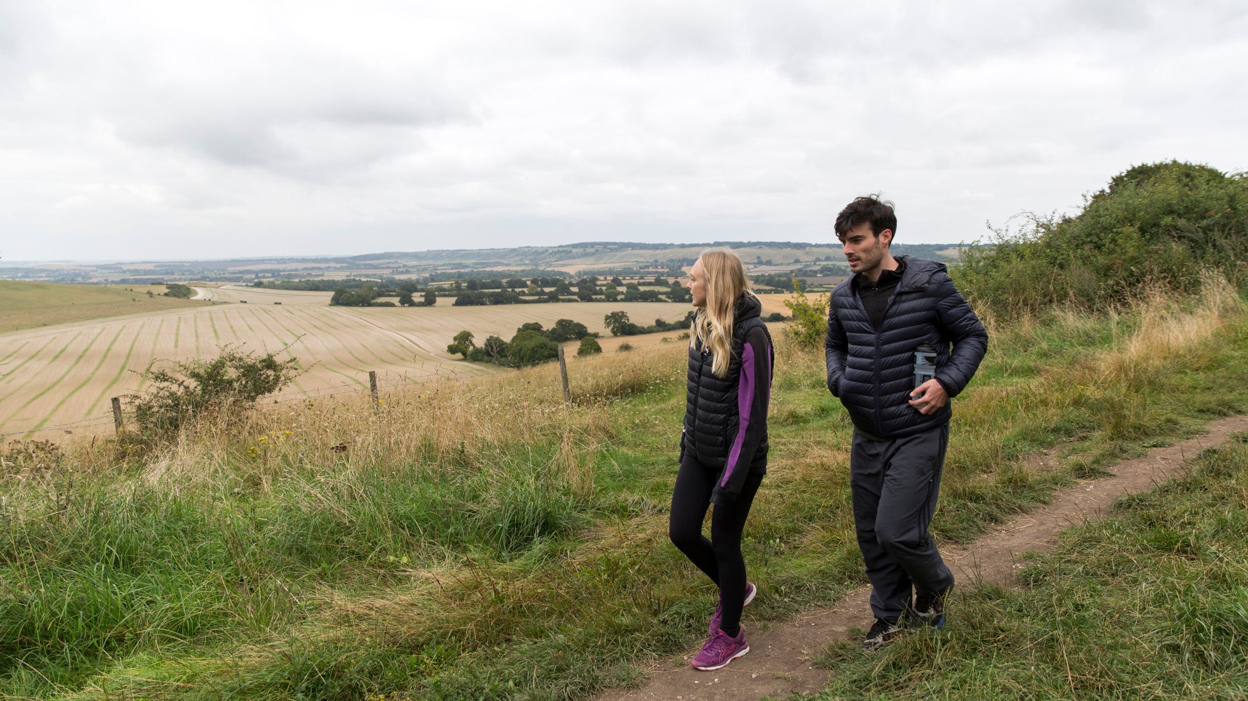 A male and female couple walking along a narrow footpath overlooking ploughed fields on a dull autumn day