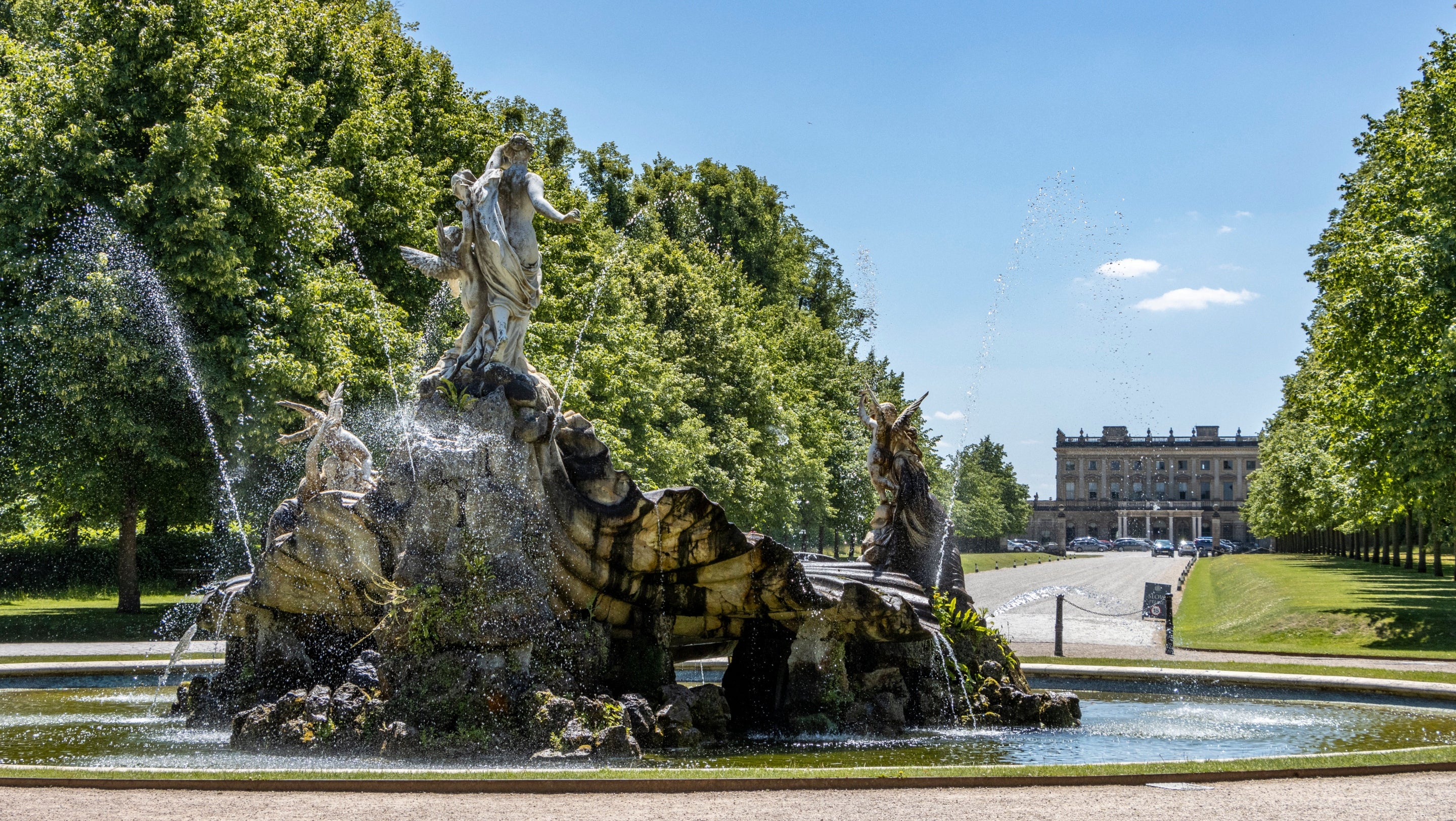 View of a large sculptural fountain in the foreground with green trees and a country house in the background with blue sky