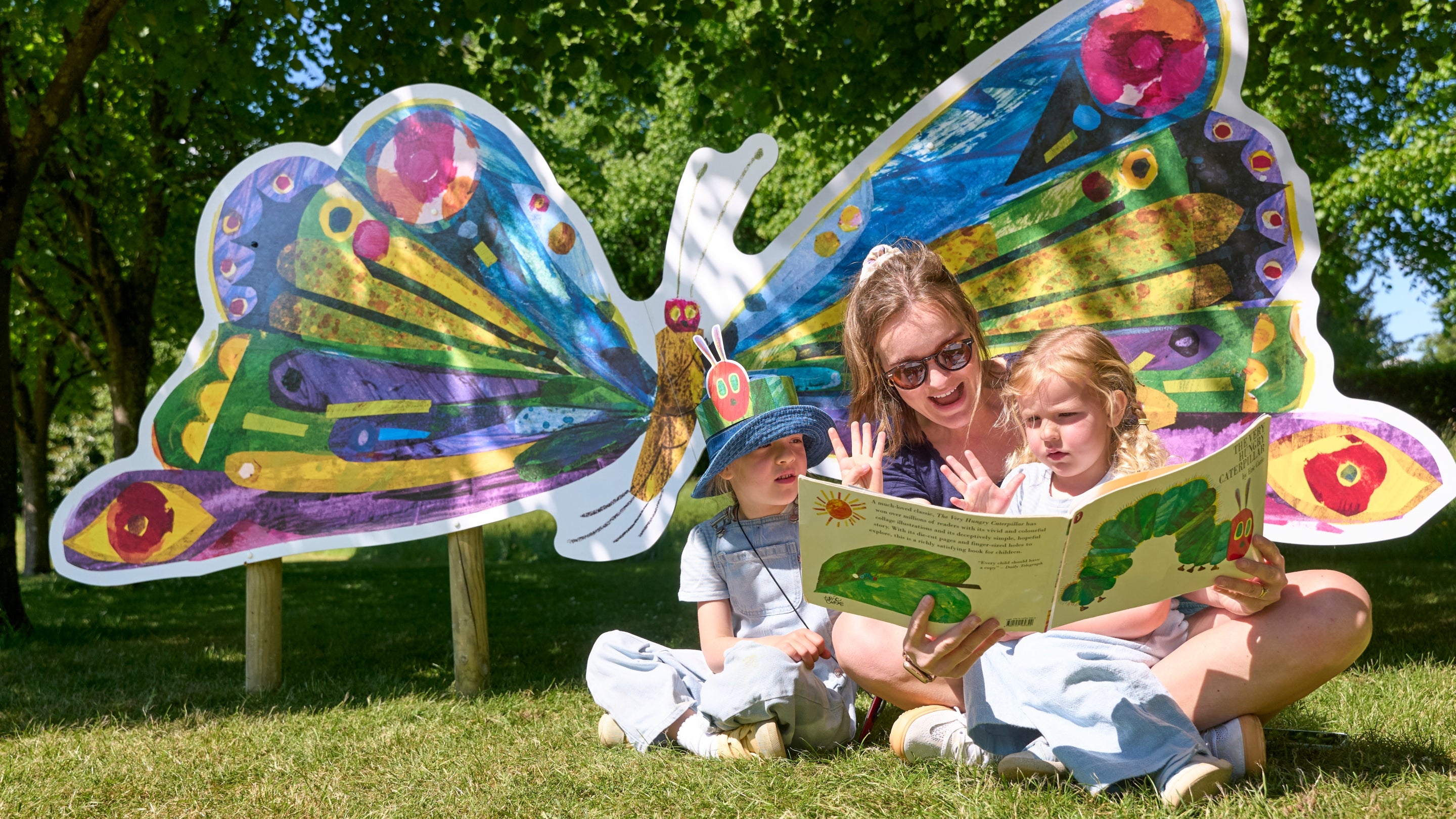 family sitting on the grass reading the Very Hungry Caterpillar book in front of a giant butterfly board