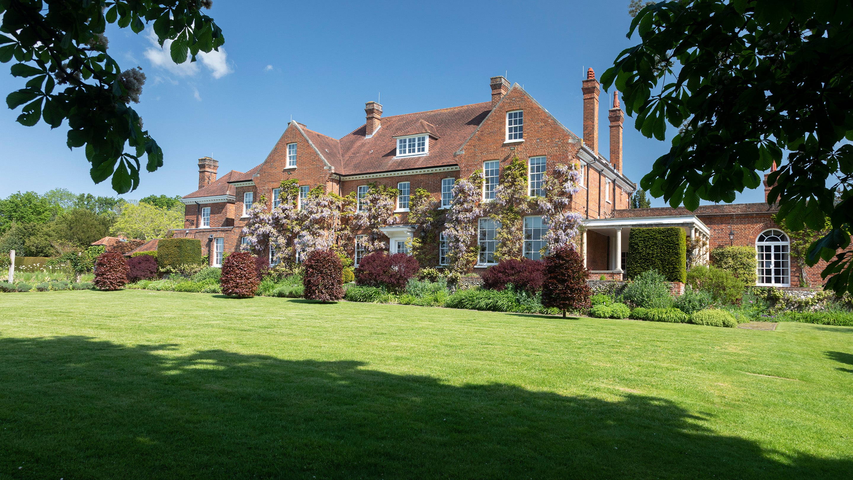 A view over well tended lawn up to the rear of a large redbrick house with 5 ornate chimneys visible.