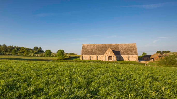 Great Coxwell Barn seen in the distance with a blue sky and vivid green grass in the foreground.