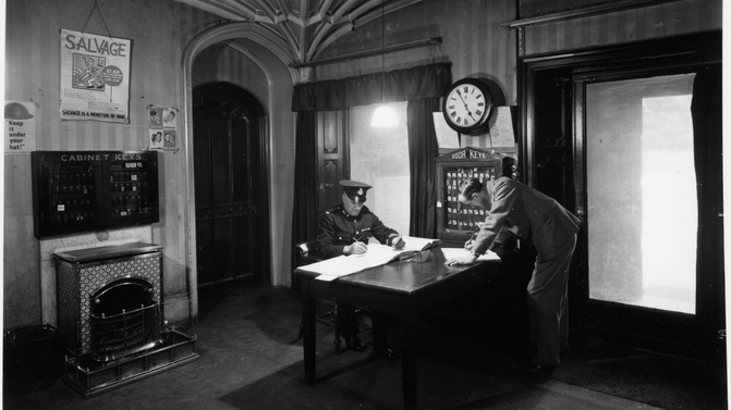 One man sat down in uniform sat at a desk with pen in hand with one man leaning over a desk also writing. Black and white photograph of the entrance hall of Hughenden Manor during Hillside.