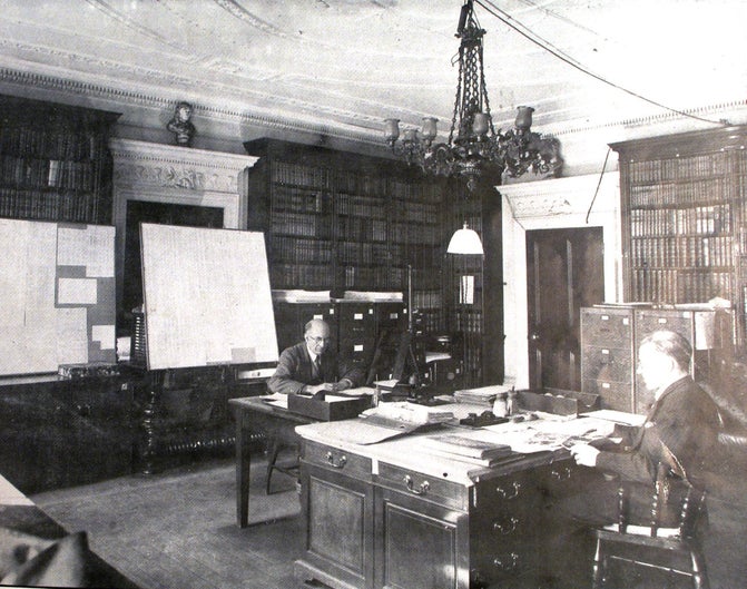 Black and white photograph of the Library at Hughenden. Two men are sat at desks surrounded by filing cabinets and pin boards.