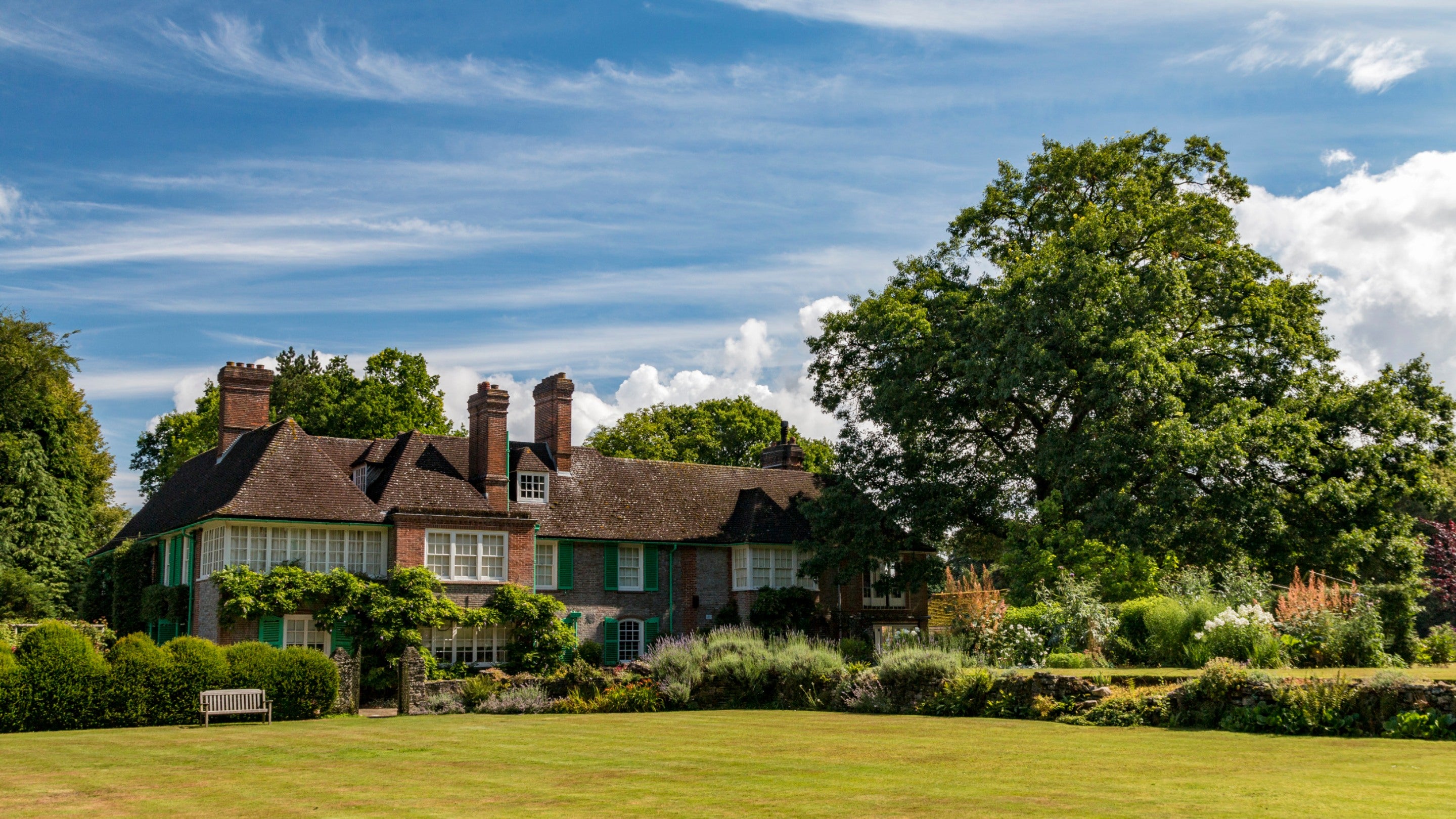 An image of the house and garden at Nuffield Place, with a neat lawn and flower borders in front of the house, a large oak tree to the right of the house and a blue sky above