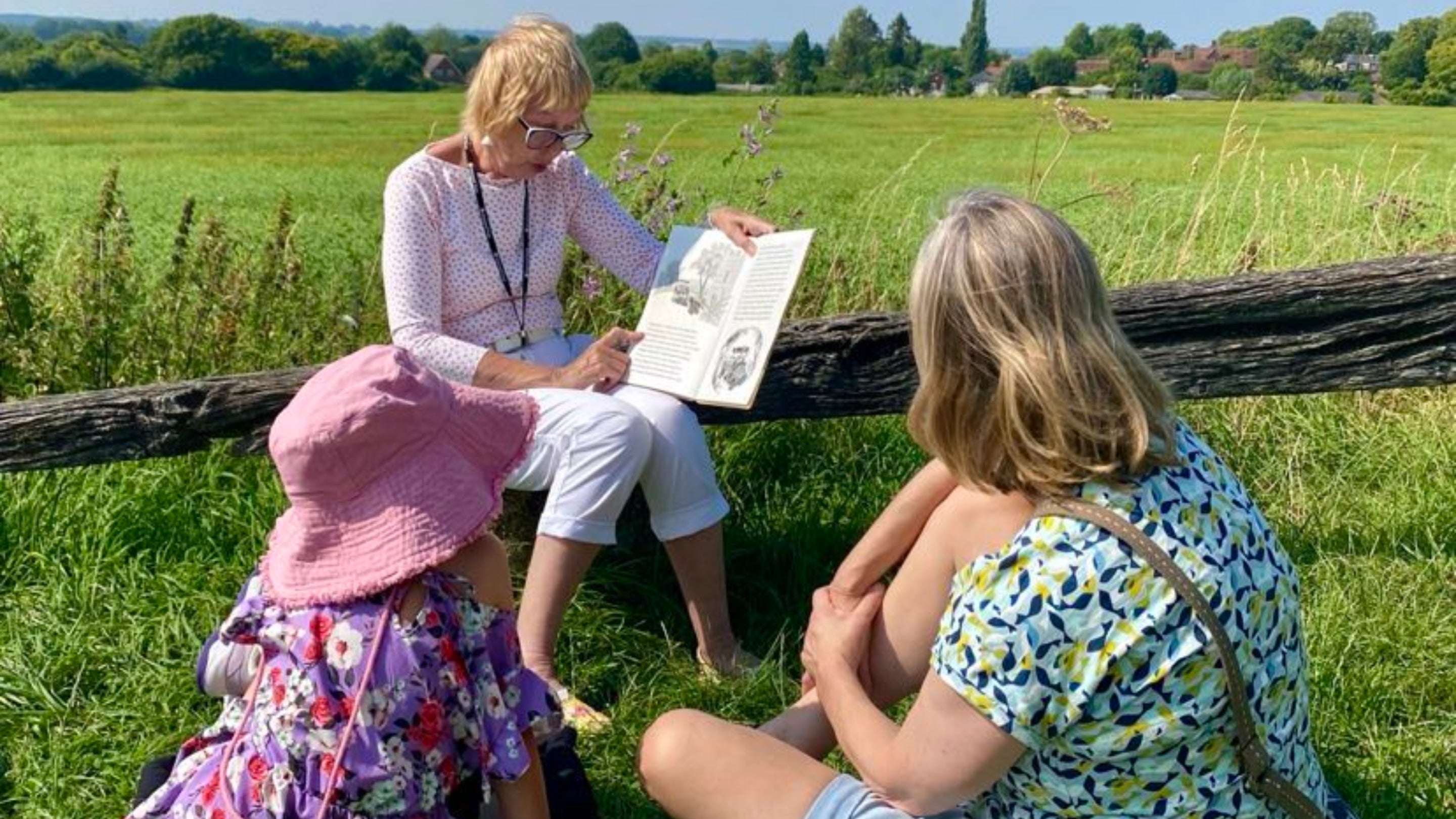 A child and an adult sit on the grass, listening to a National Trust Volunteer reading from a picture book at Pitstone Windmill.