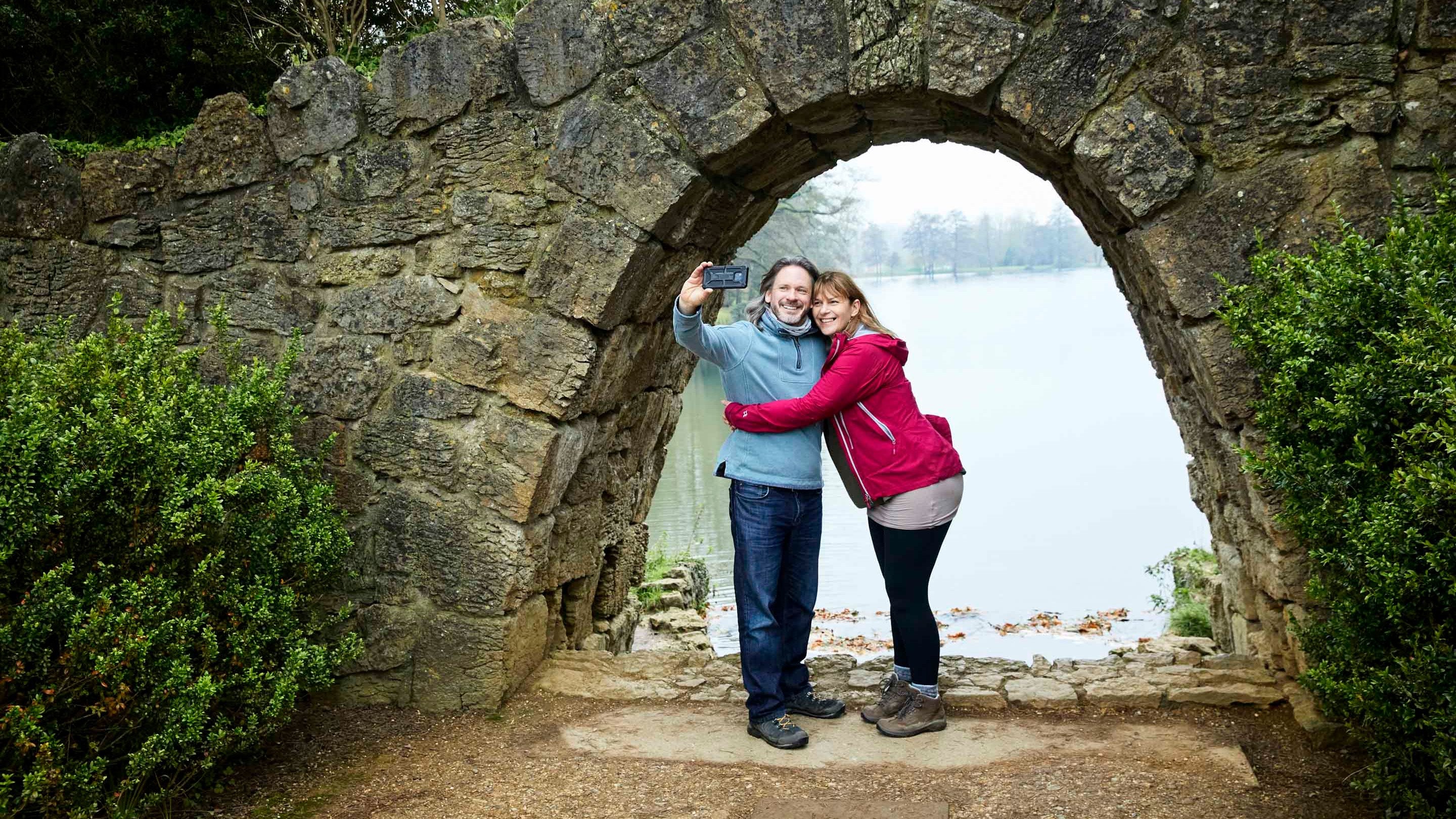 A couple stand in front of the arch of the cascade taking a selfie with the Eleven Acre Lake behind them at Stowe, Buckinghamshire.