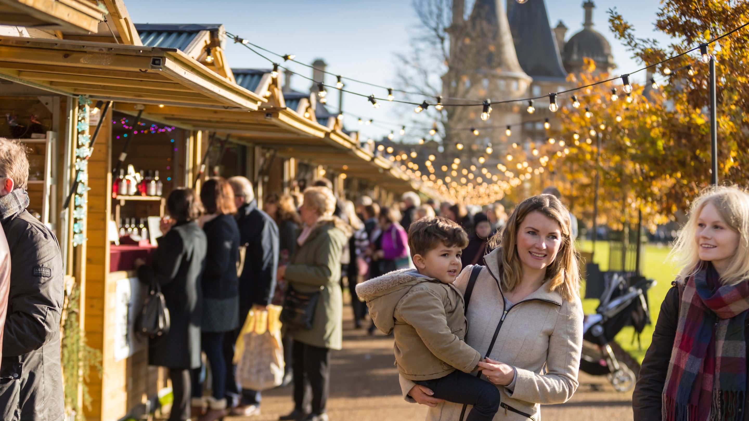 Two women, one holding a small child, stand by the wooden stalls and fairy lights of the Christmas market, busy with shoppers, at Waddesdon Manor, Buckinghamshire