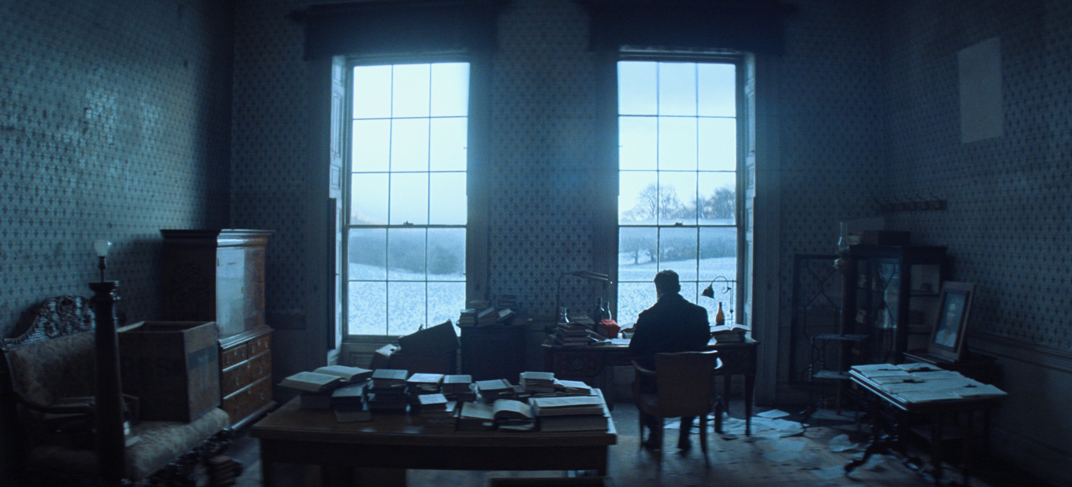 A man sits at a writing desk, facing a window that overlooks the parkland