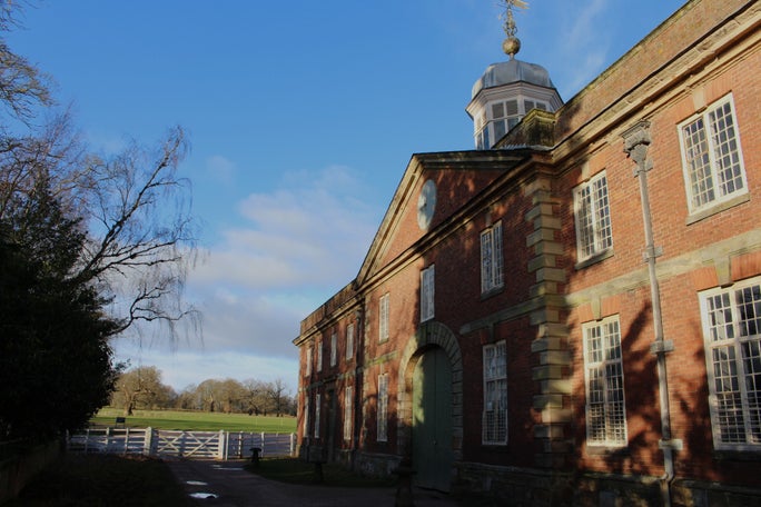 Beside the stableyards at Calke Abbey, with a large redbrick building and a blue bell tower to the right and white gates set into a fence at the end of the path. Green grass and tarmac are visible beyond them.