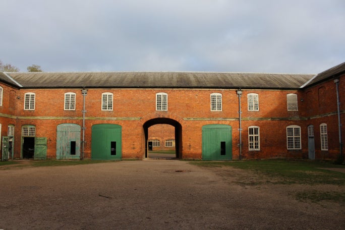Red brick buildings in the stableyards at Calke Abbey surround a large courtyard. An archway in the centre leads through to another courtyard in the distance, with bright green painted doors framing either side.