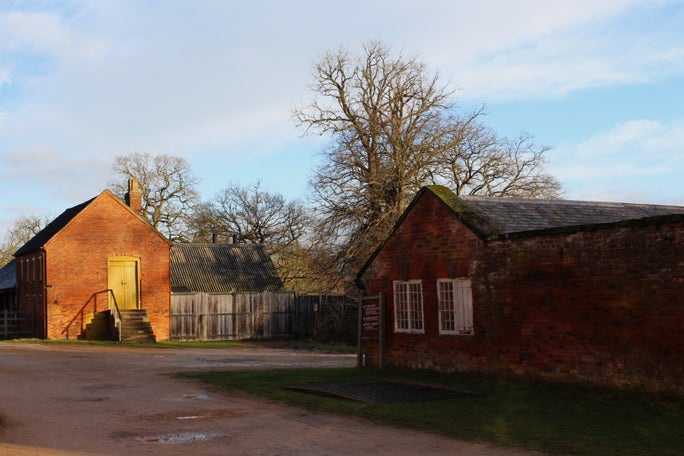 The Weighbridge at Calke Abbey, which sits in grass beside a large gravel pathway. Behind it are multiple redbrick outbuildings, and trees are visible against blue skies.