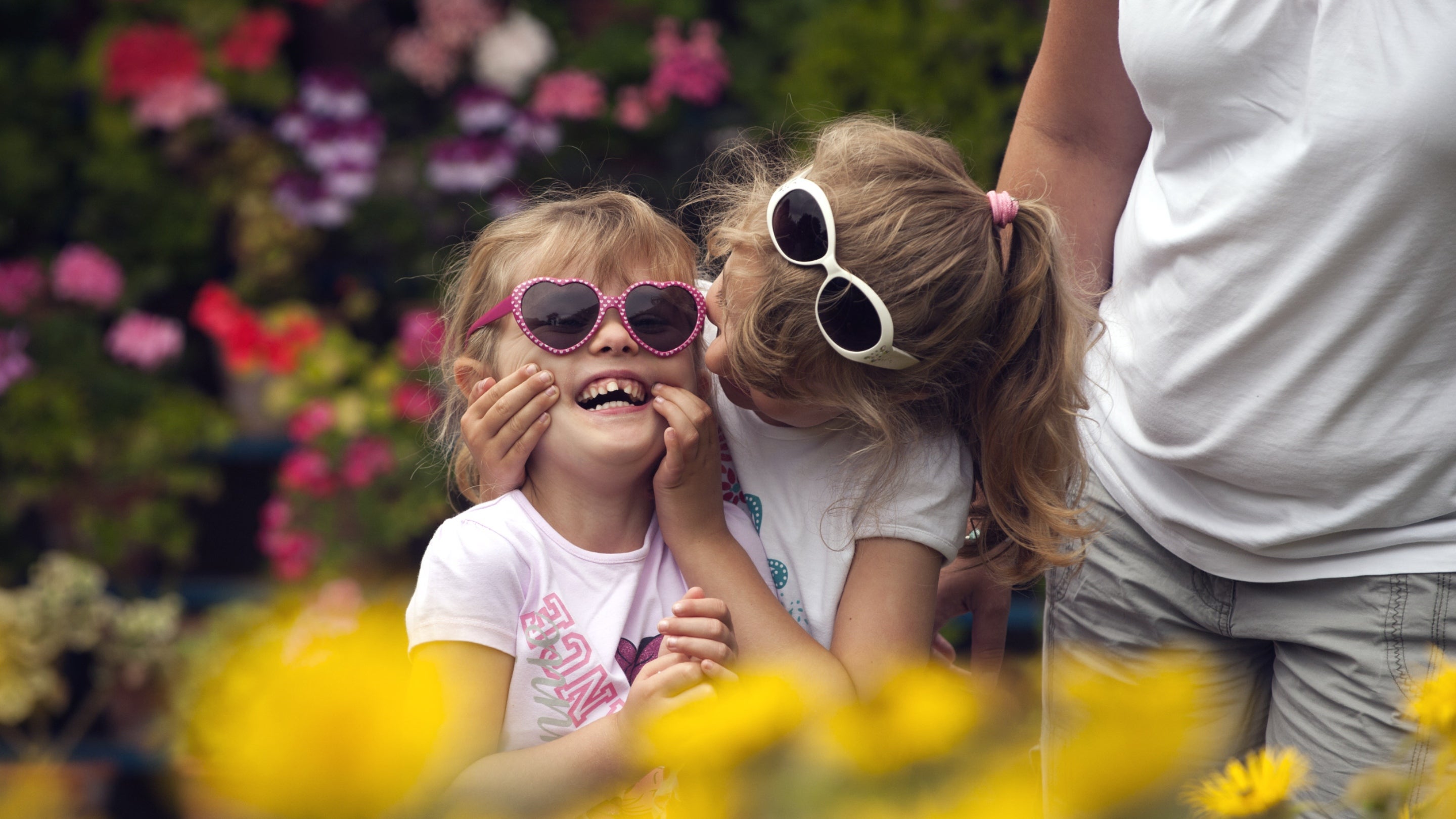 A child wearing heart-shaped sunglasses laughing as another child cups their cheeks, with yellow flowers in the foreground and lots of colourful flowers visible in the background at Calke Abbey garden
