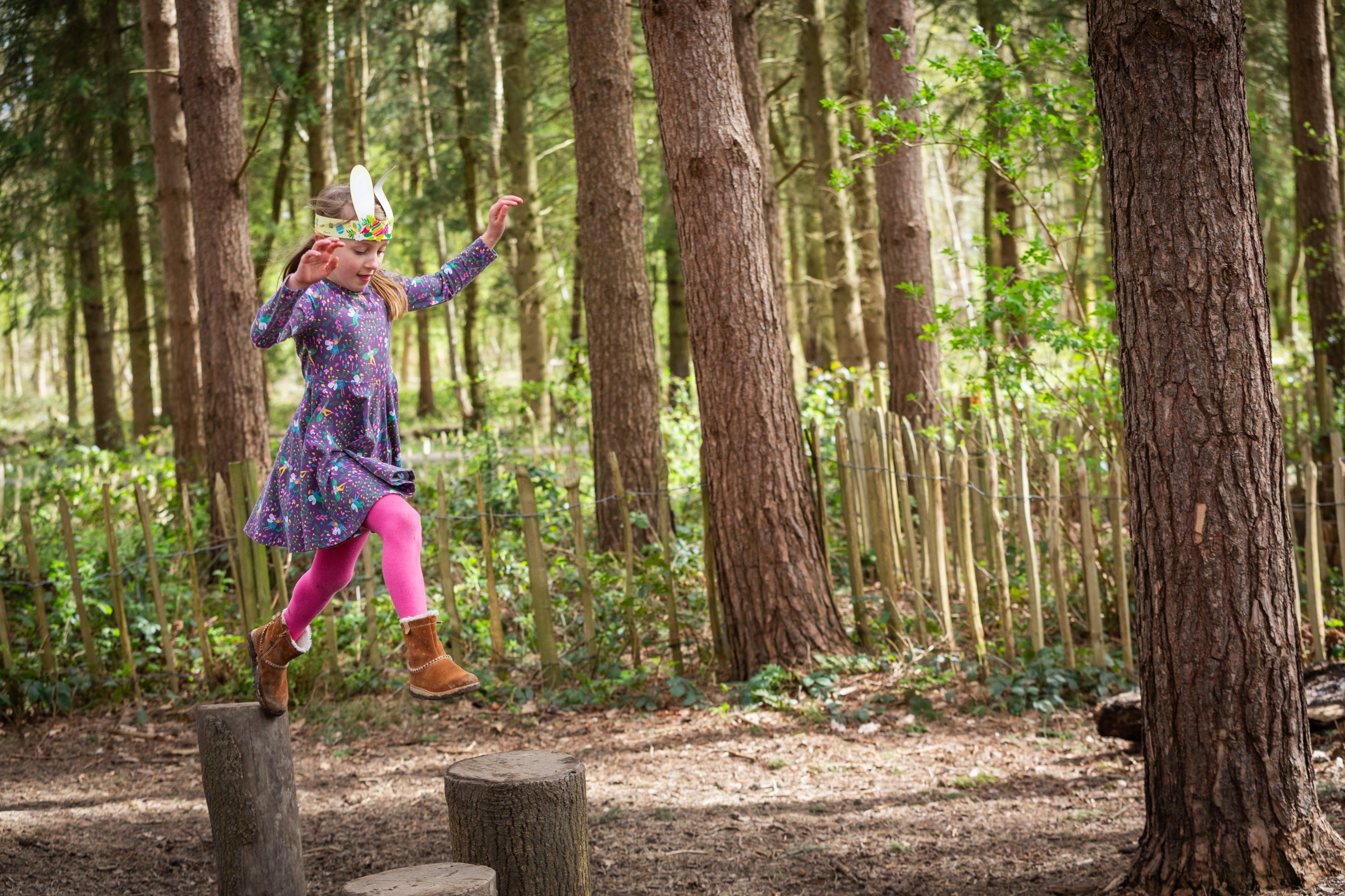 A young girl wearing Easter bunny ears leaps across wooden logs at the natural play area at Calke Explore