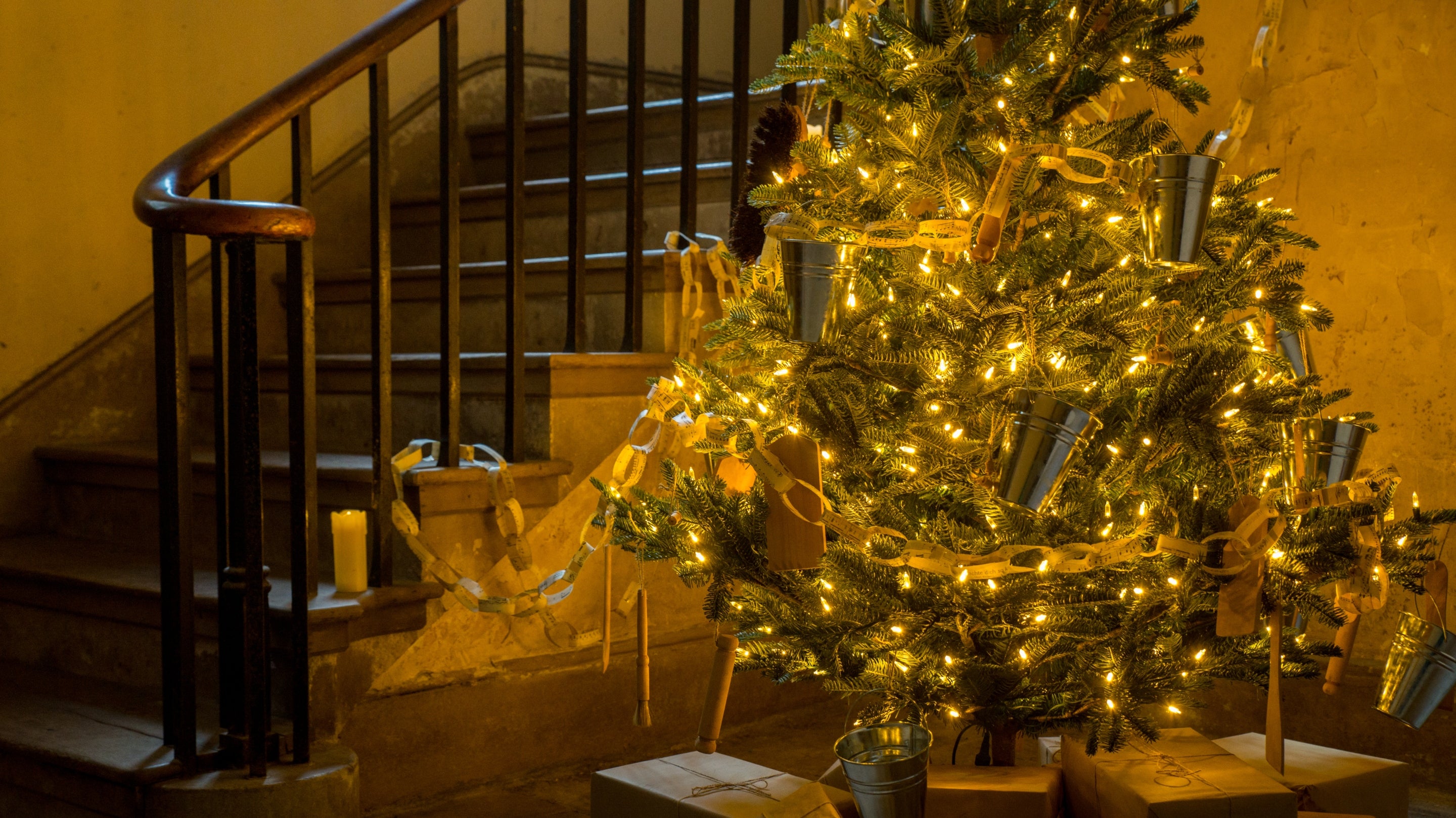 A tree decorated with bright lights and paper chains, with presents wrapped in brown paper beneath it, at the base of a staircase in the house at Calke Abbey.