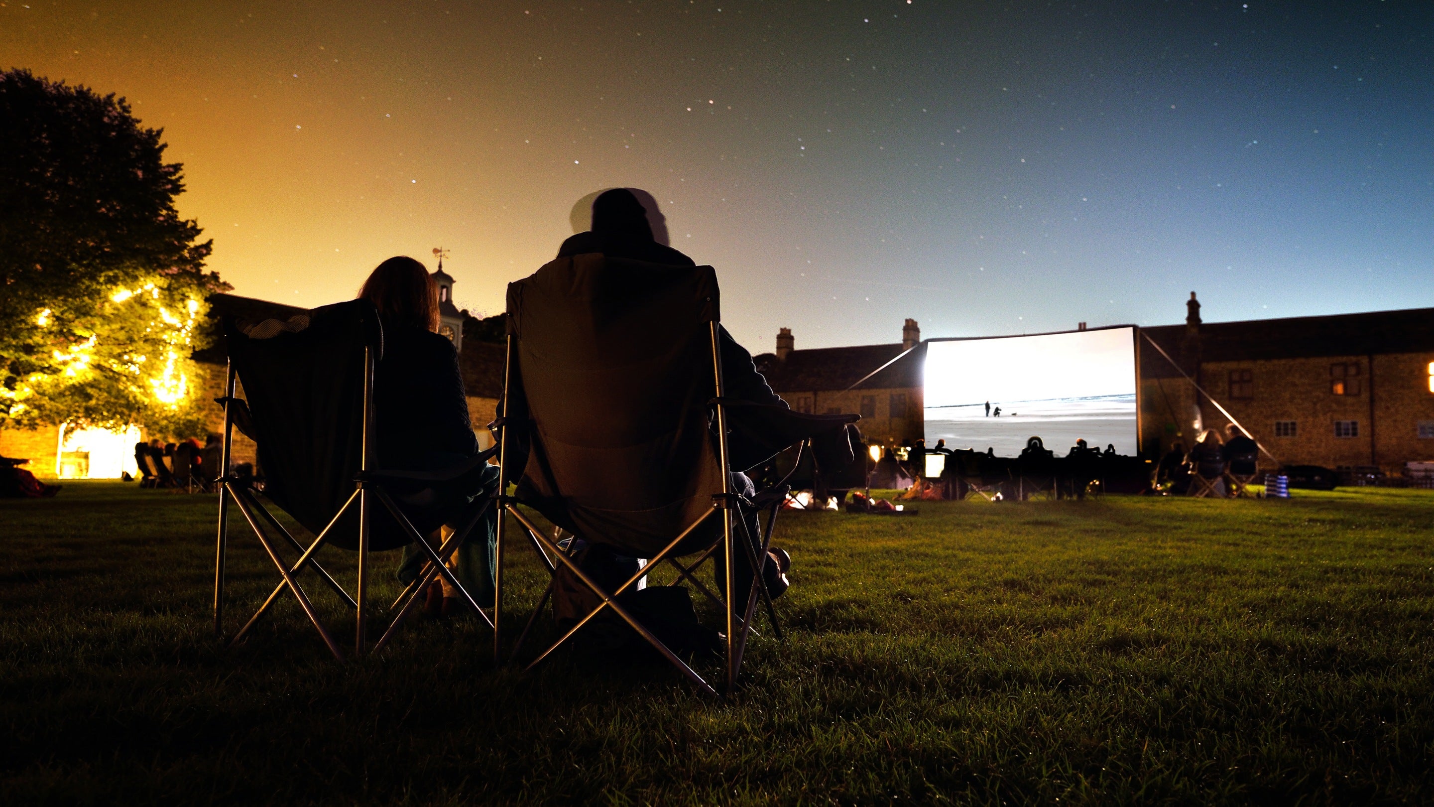Two visitors sitting in folding chairs at dusk with a film showing on an outdoor screen at a cinema experience at Hardwick, Derbyshire.
