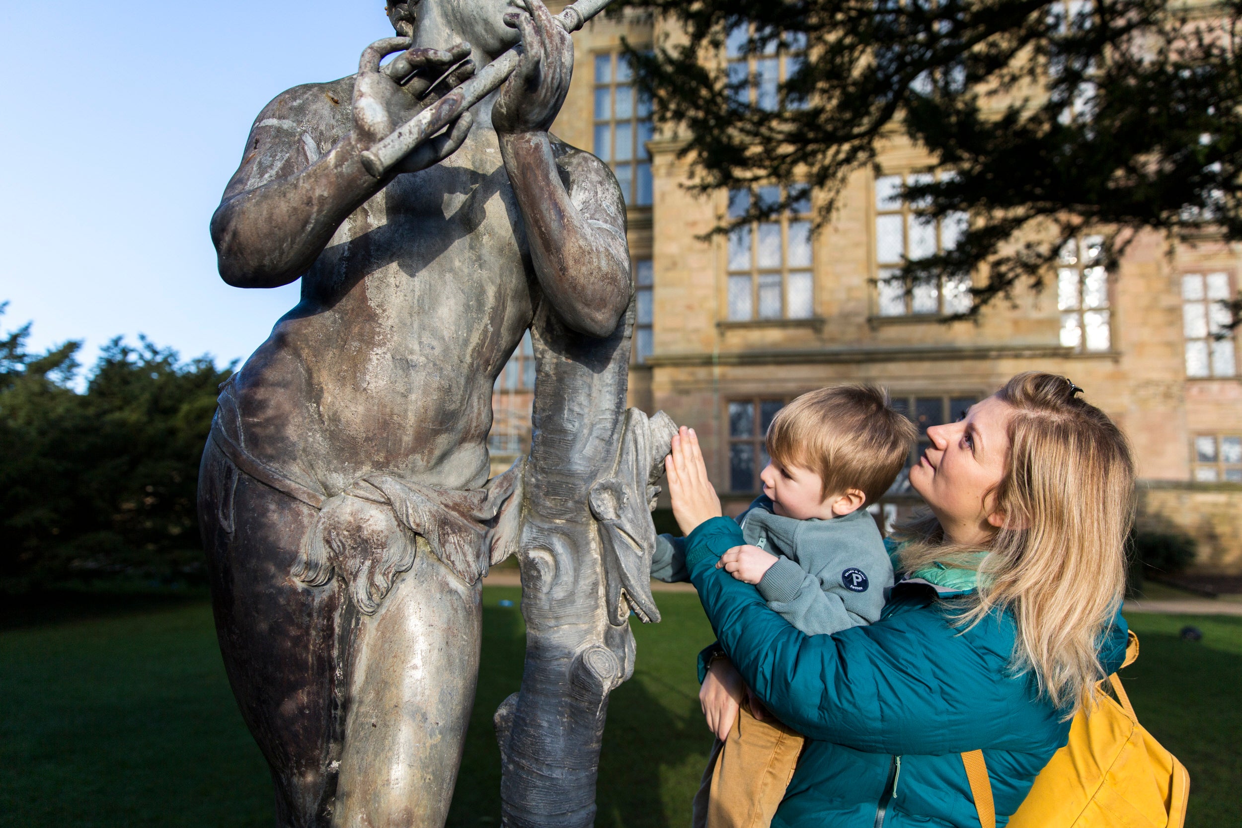 A mother with a teal coat and bright yellow backpack, holding her young son with light brown hair, reaches for a grey statue in the gardens, with the sand-coloured Hardwick Hall seen in the backdrop.