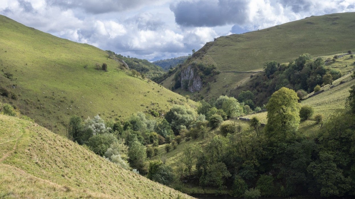 Looking down a valley with grasslands and woodlands on the slopes and valley bottoms with a rocky cliff face in the distance under grey dramatic skies.