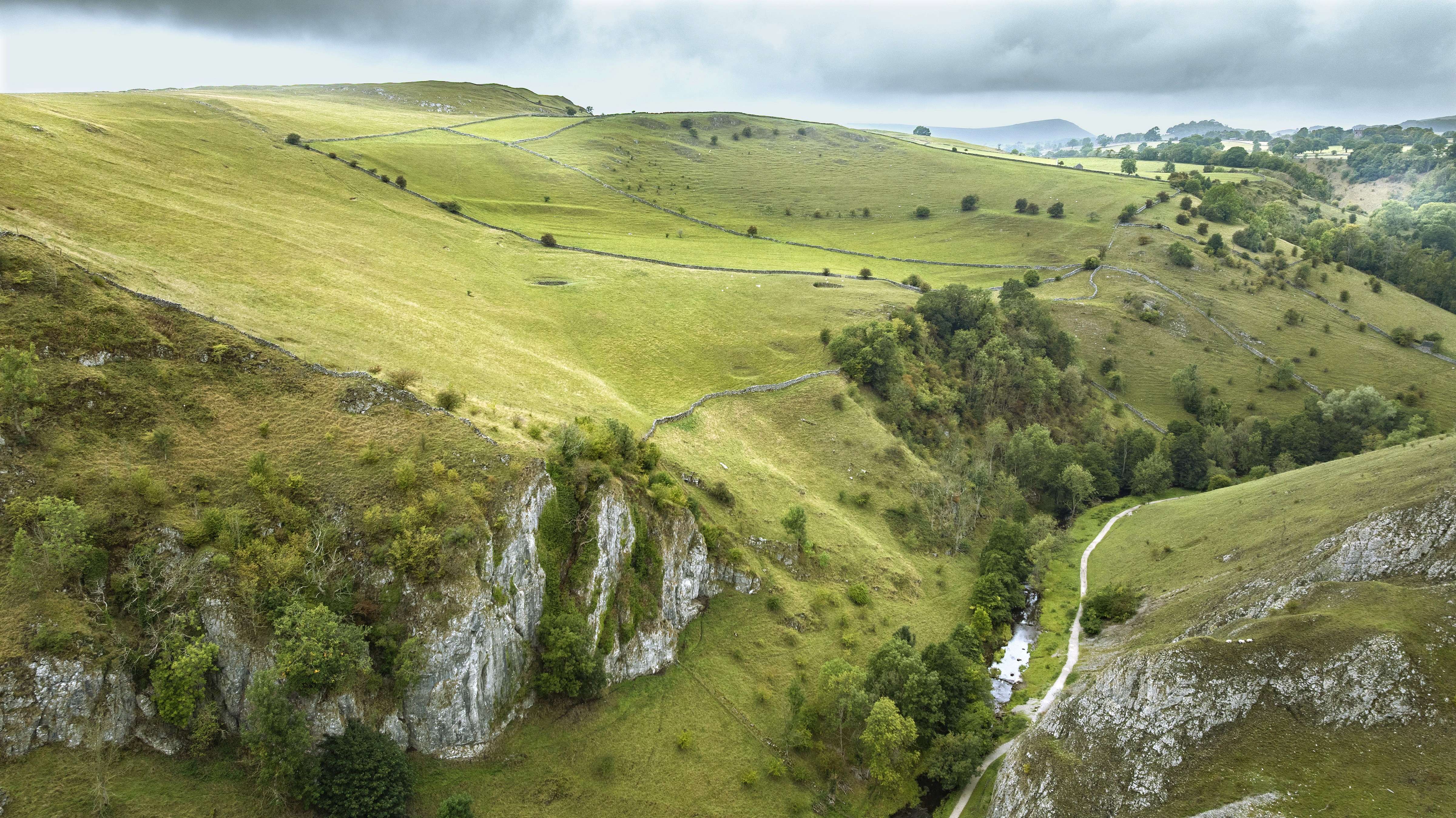 An aerial view looking down on fields on the hills on top of limestone rock faces above a valley with a river running through it. There are some trees in the valley and up the sides of the ravines. Drystone walls divide the fields.