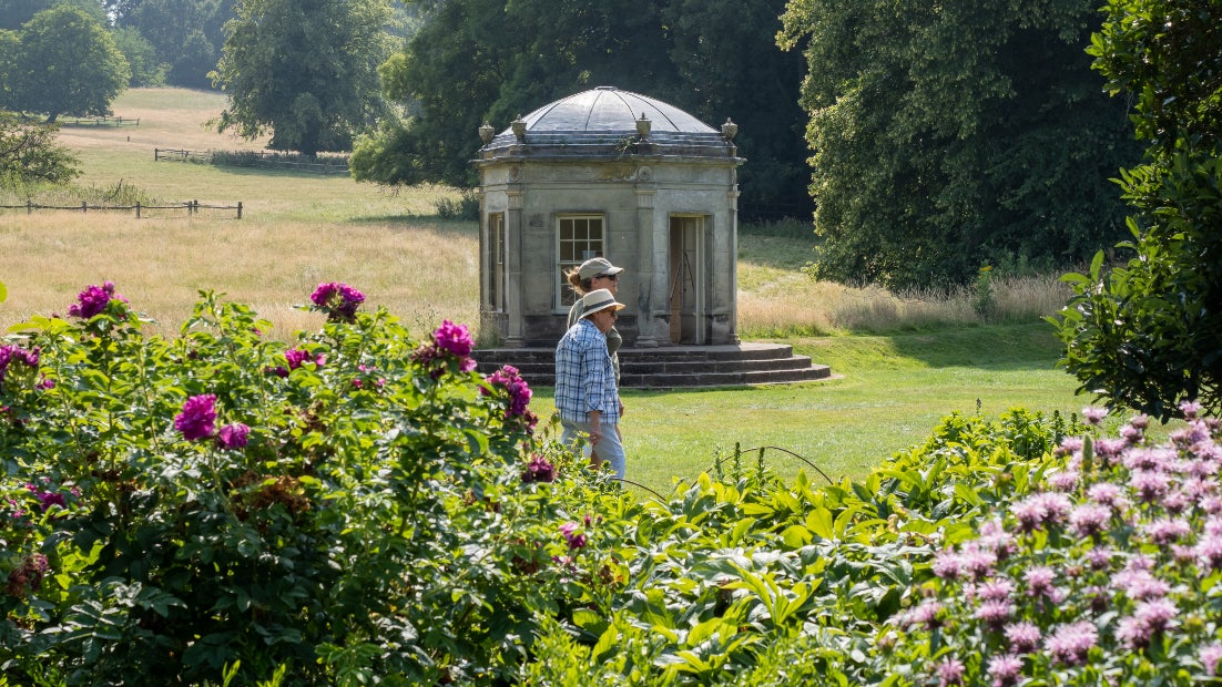 Two adults walking across Kedleston Hall's garden with a summer house in the background and flower beds in the foreground