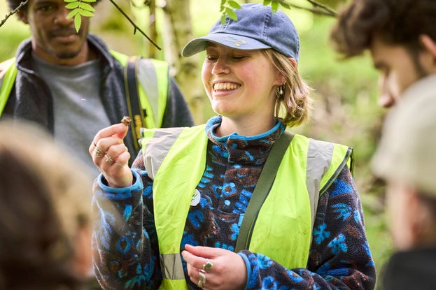 A group of people stood around a person wearing a baseball cap and a hi-vis jacket who is smiling as they look at a plant.