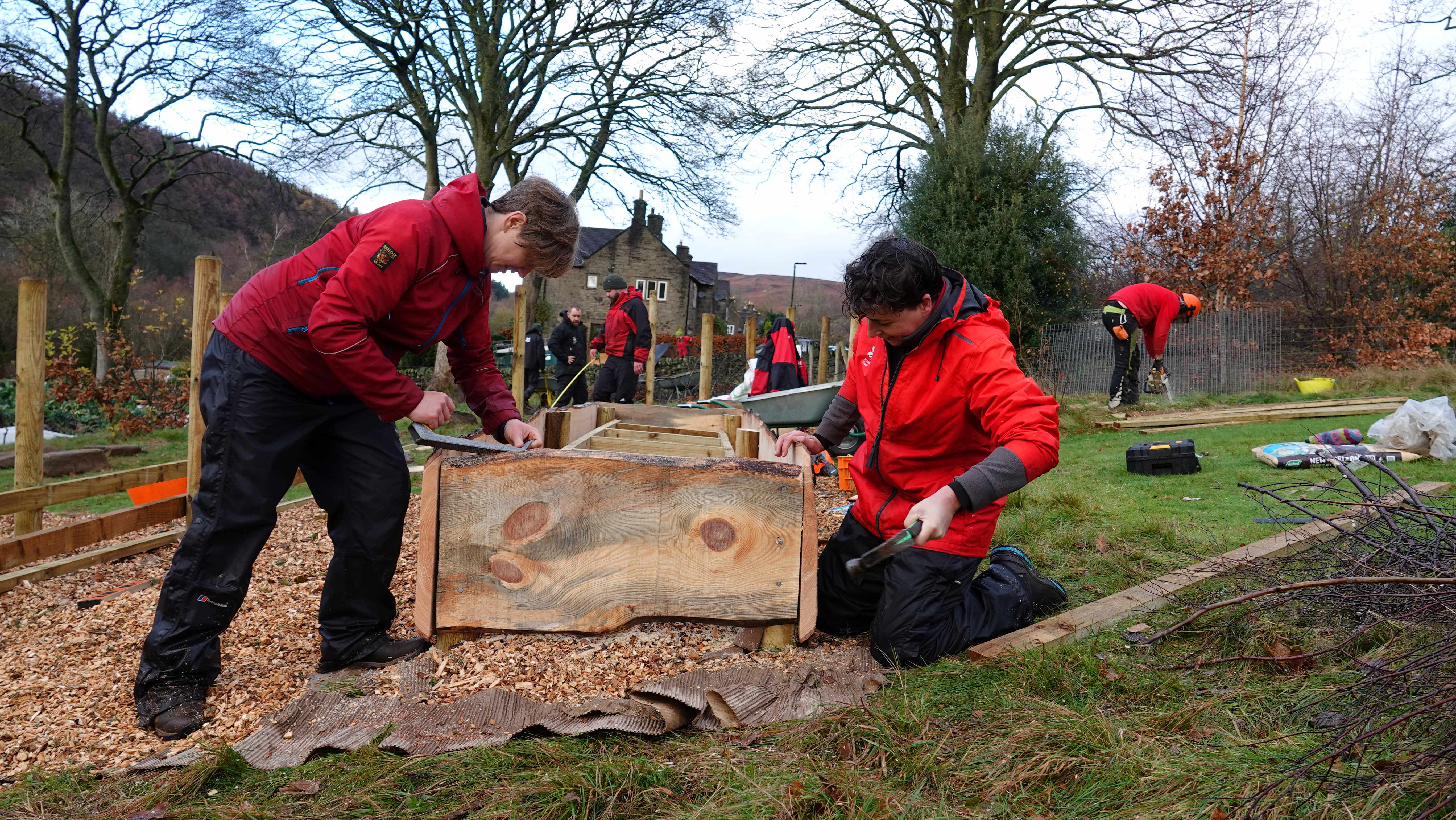 Two people in the foreground using tools to build the frame for a raised bed with other rangers and volunteers working in the background.